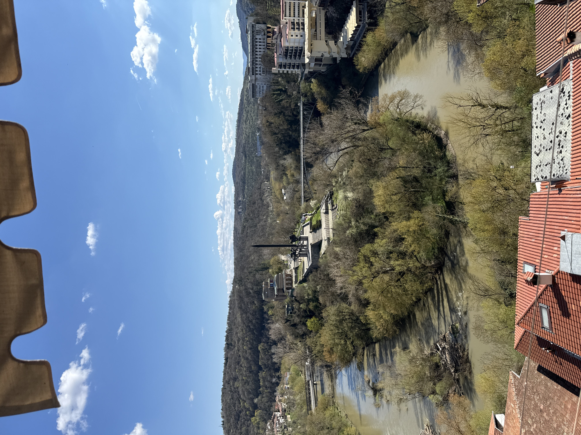 The image depicts a high-angle view in Veliko Tarnovo, Bulgaria. The Yantra River meanders through the scene, with dense vegetation along its banks. On the left side of the river, a wooded hillside rises, culminating in a monument with a tall, dark obelisk. Further up the river on the opposite bank are several multi-story buildings. A bridge spans the river in the middle of the image. In the foreground, on the right, are the tiled roofs and a facade of a building. The sky is blue with scattered white clouds. Part of a structure is visible in the upper left corner.