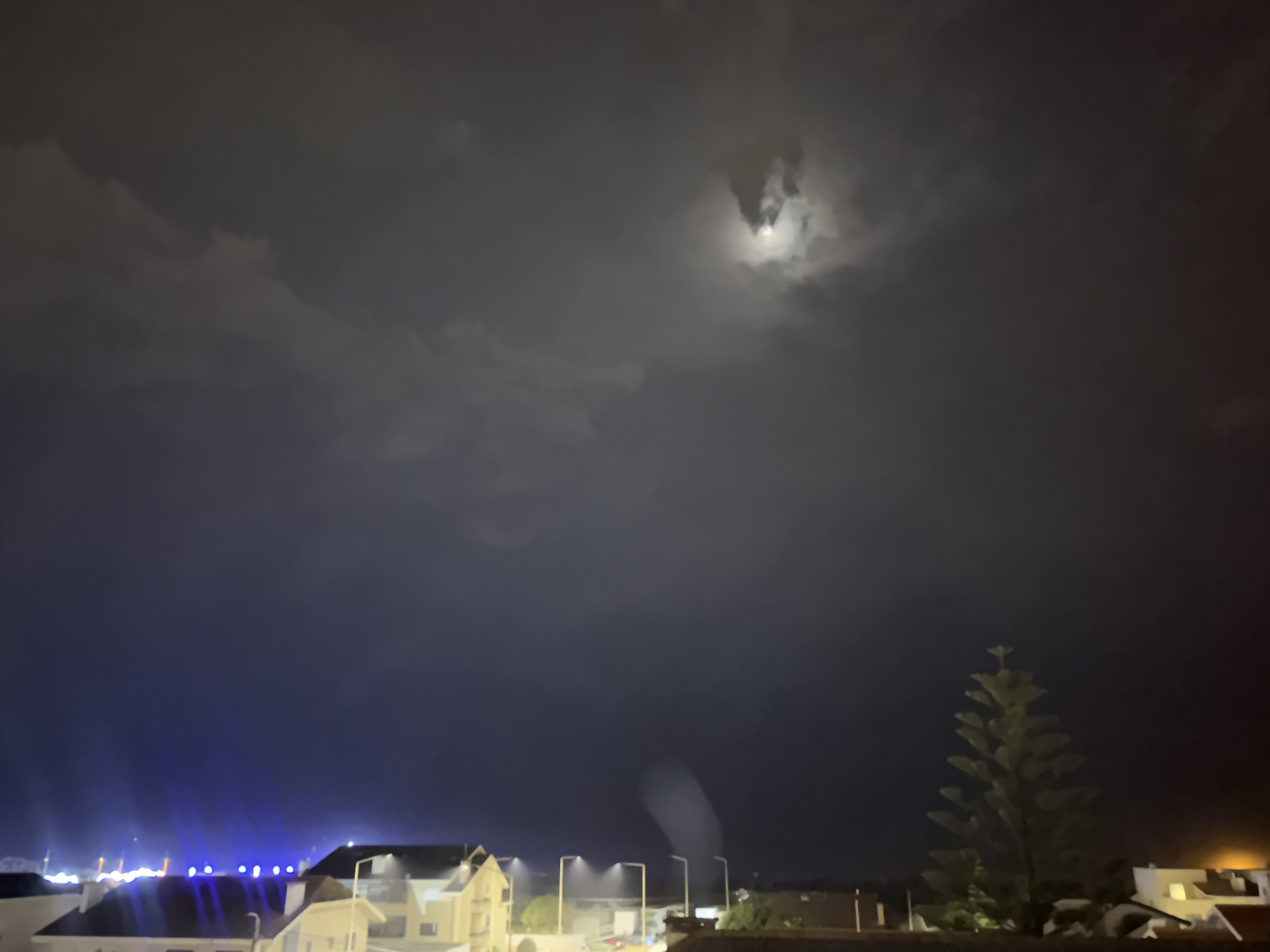 The image depicts a nighttime scene in Vila Nova de Gaia, Portugal. The sky is heavily clouded, with a visible moon partially obscured by the clouds. Below, there are buildings, streetlights, and what appears to be a tree silhouette on the right side of the frame. In the lower-left corner, there are blue lights and building rooftops. The foreground is obscured by a dark fence. The image is taken from a slightly elevated position. A column of vapor ascends from the lower-center part of the scene.