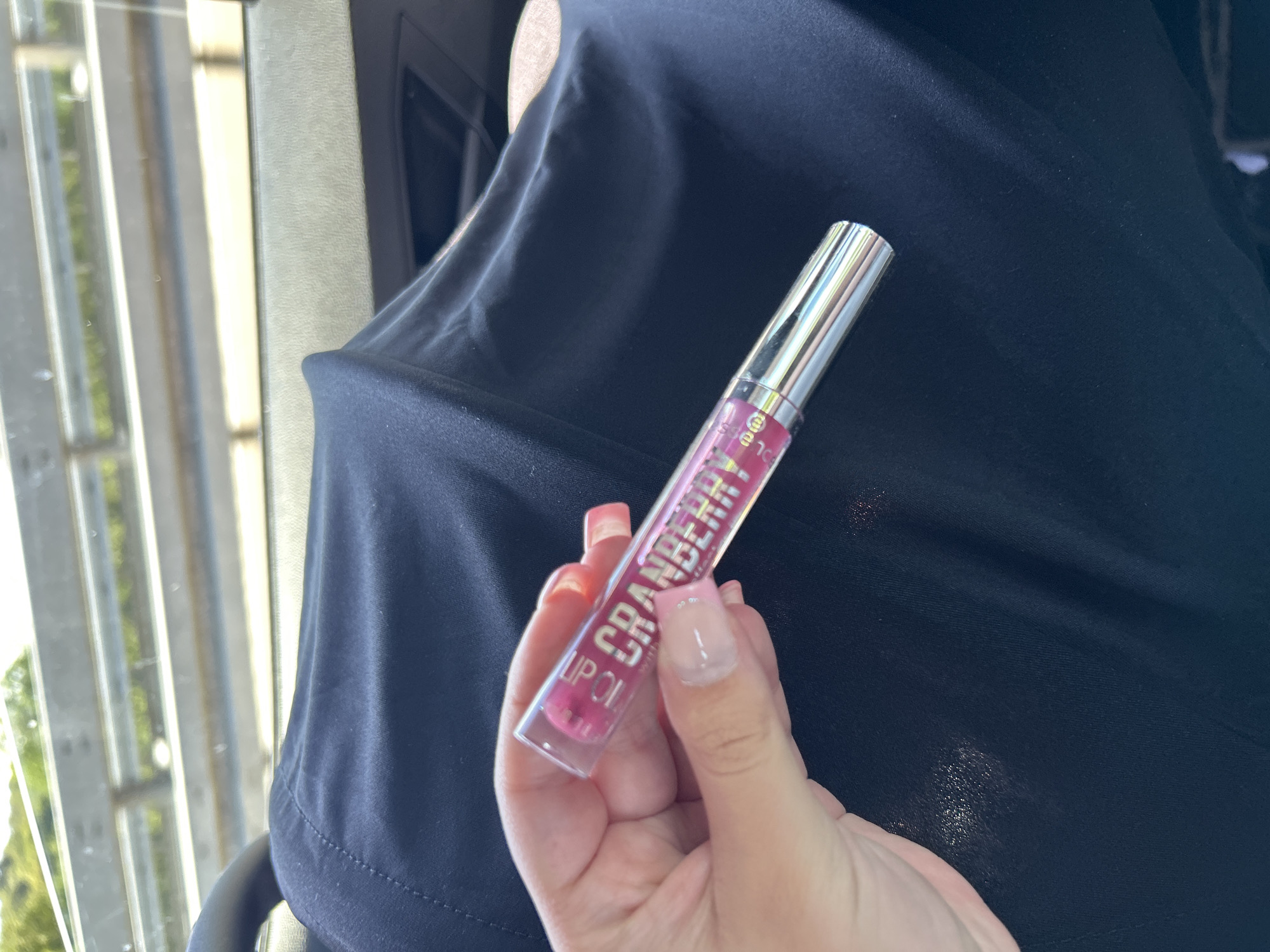 The image shows a close-up of a hand holding a tube of lip gloss. The lip gloss tube is clear with pink lettering that reads "LIP OIL CRANBERRY". The hand has long, artificial nails with a light pink color. The background is out of focus, but appears to be the interior of a vehicle, with a dark fabric material and a blurred view of greenery and a railing through a window. The scene is likely in Reichshof, Germany, based on the provided location information. The lighting suggests it is daytime.