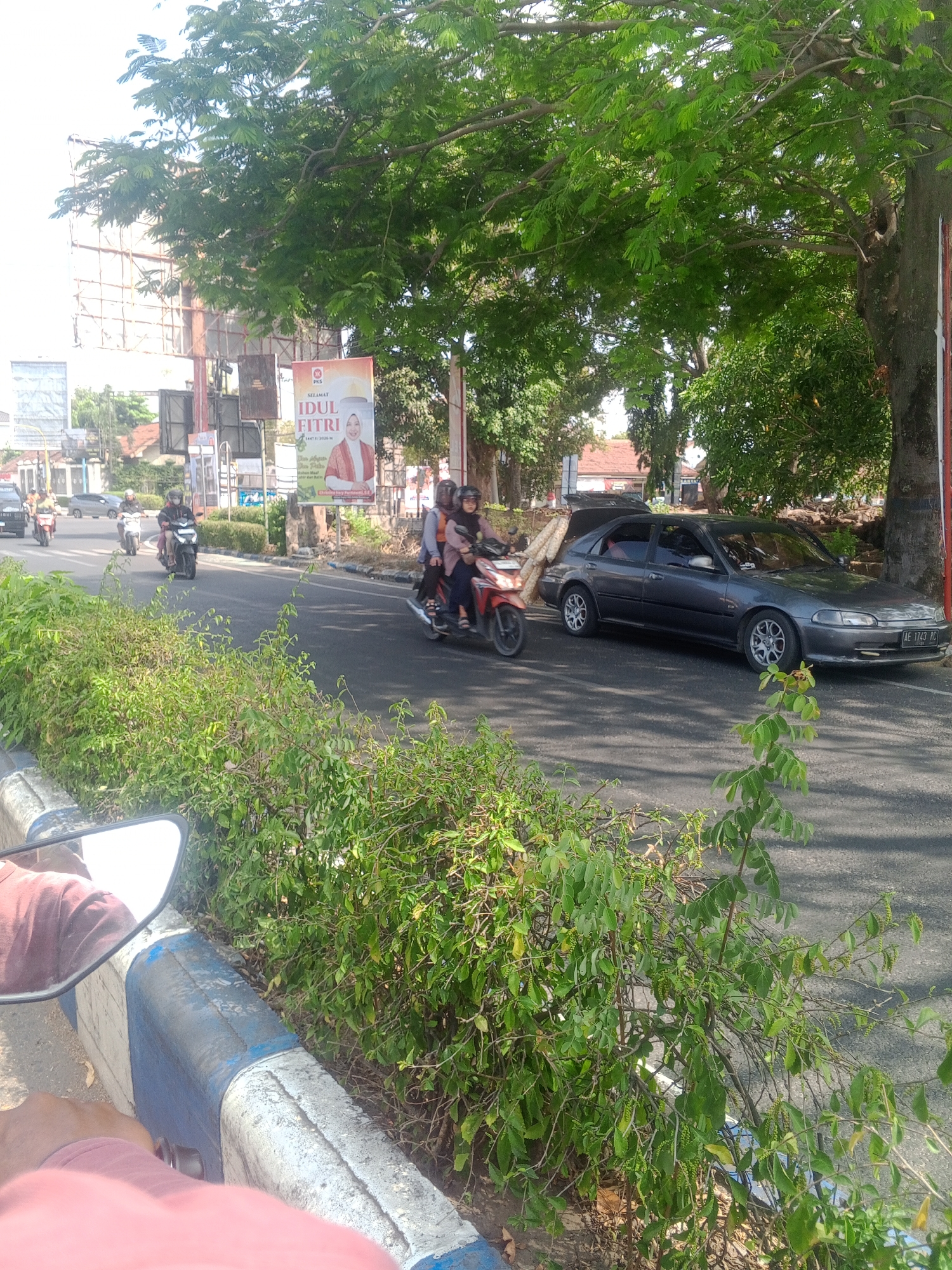 The image depicts a street scene in Tonatan, Indonesia, likely during the day, given the bright lighting and shadows. In the foreground, a person's arm, wearing a pink shirt, is visible, along with a motorcycle's rearview mirror reflecting the sky and trees. The road is paved, with a blue and white concrete barrier running along its edge, lined with lush green bushes.

Further down the road, a dark gray sedan is parked on the right side, its trunk open and filled with what appears to be wooden poles. To the left of the sedan, a motorcycle with two occupants, a driver and a passenger wearing hijabs, is in motion. The driver is wearing a dark jacket and helmet, while the passenger has on an orange top and a hijab.

Several other motorcycles are also seen on the road, with riders wearing helmets and casual clothing. Some are moving forward, while one is stationary. In the background, a large billboard displays a message that reads "SELAMAT IDUL FITRI" (Happy Eid al-Fitr) with an image of a woman in traditional attire. Behind the billboard, buildings and trees are visible, contributing to the urban and natural mix of the environment. The overall scene suggests a typical day in a residential or commercial area of Tonatan, with a sense of everyday activity and movement.