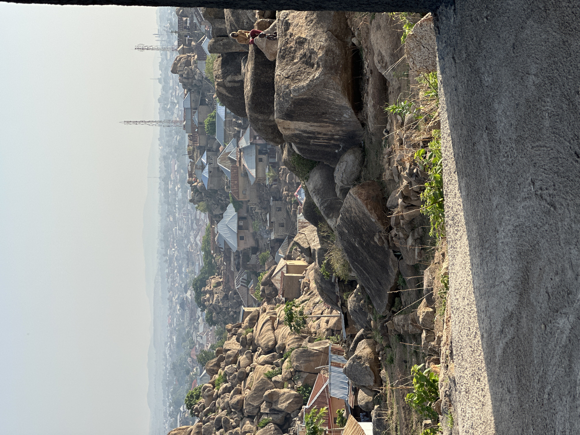 The image depicts a cityscape with buildings nestled among large rock formations, viewed from an elevated position. The location is likely within the city of Jos, Nigeria, given its known geology and urban development integrated with rocky terrain. In the distance, the city extends into a hazy horizon, with telecommunication towers visible. In the middle ground, tin-roofed houses are scattered amongst the boulders. At the top of the rocks in the middle ground, there are two human figures, one is dressed in khaki, the other is wearing red. The foreground includes the edge of a grey concrete structure from where the image is captured. Vegetation grows among the rocks, closer to the viewpoint.