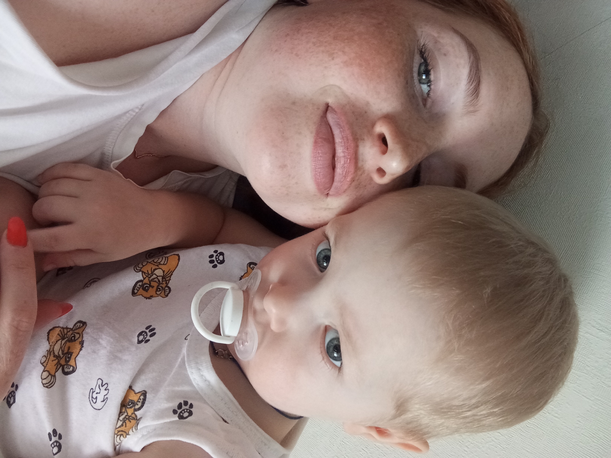 The image shows a woman and a baby. The woman has fair skin, freckles, and light blue eyes. Her lips are pale pink. She is wearing a white shirt. The baby is lying against her. The baby has fair hair, light blue eyes, and a pacifier in their mouth. The baby is wearing a white shirt printed with Simba (Lion King) images and paw prints. A woman's hand with red nail polish is visible in the lower left. The background is light gray. There is no explicit information about the location in Yurgamysh, Russia.