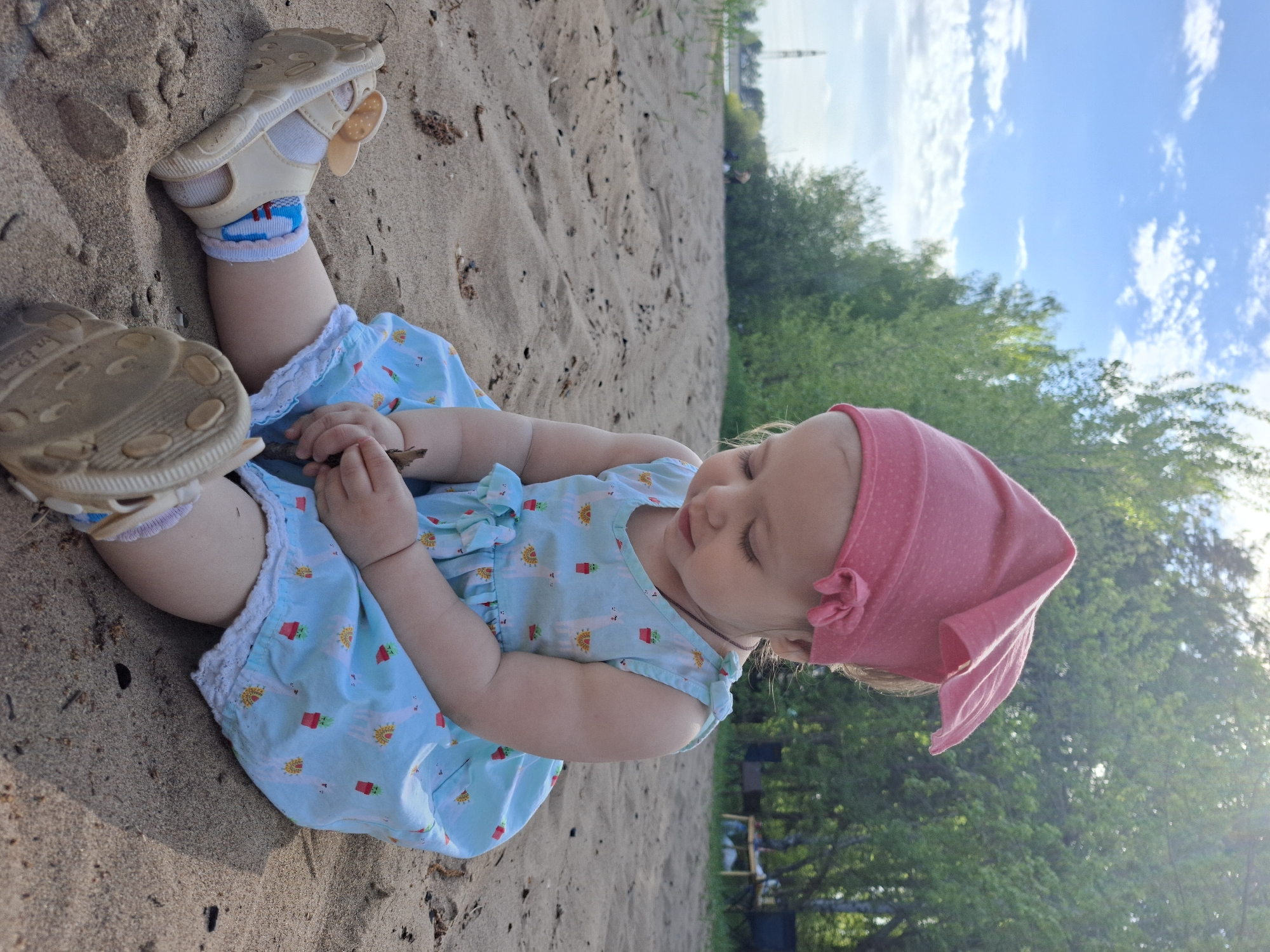 A young child sits on a sandy surface, likely a beach, wearing a blue dress with a cactus pattern and a pink headscarf. The child is holding a small stick or piece of wood in their hands. Their sandals are visible nearby. The background shows greenery and a glimpse of the sky, and a slender, light-colored structure (possibly a tower or tall building) in the distance. The location appears to be in Noviy, Russia.