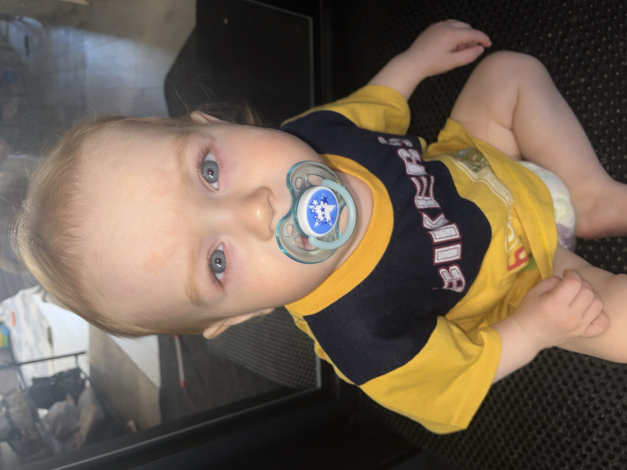 A close-up, high-angle shot depicts a baby lying on what appears to be a dark, textured surface. The baby has fair skin, blonde hair, and blue eyes, with a pacifier in their mouth. They are wearing a yellow shirt with navy blue accents and a design with white letters. The baby also seems to be wearing a diaper. Part of a reflective surface (possibly glass) can be seen to the left, with indistinct shapes reflected in it. The location of Chaikovsky, Russia is not discernible from the image.