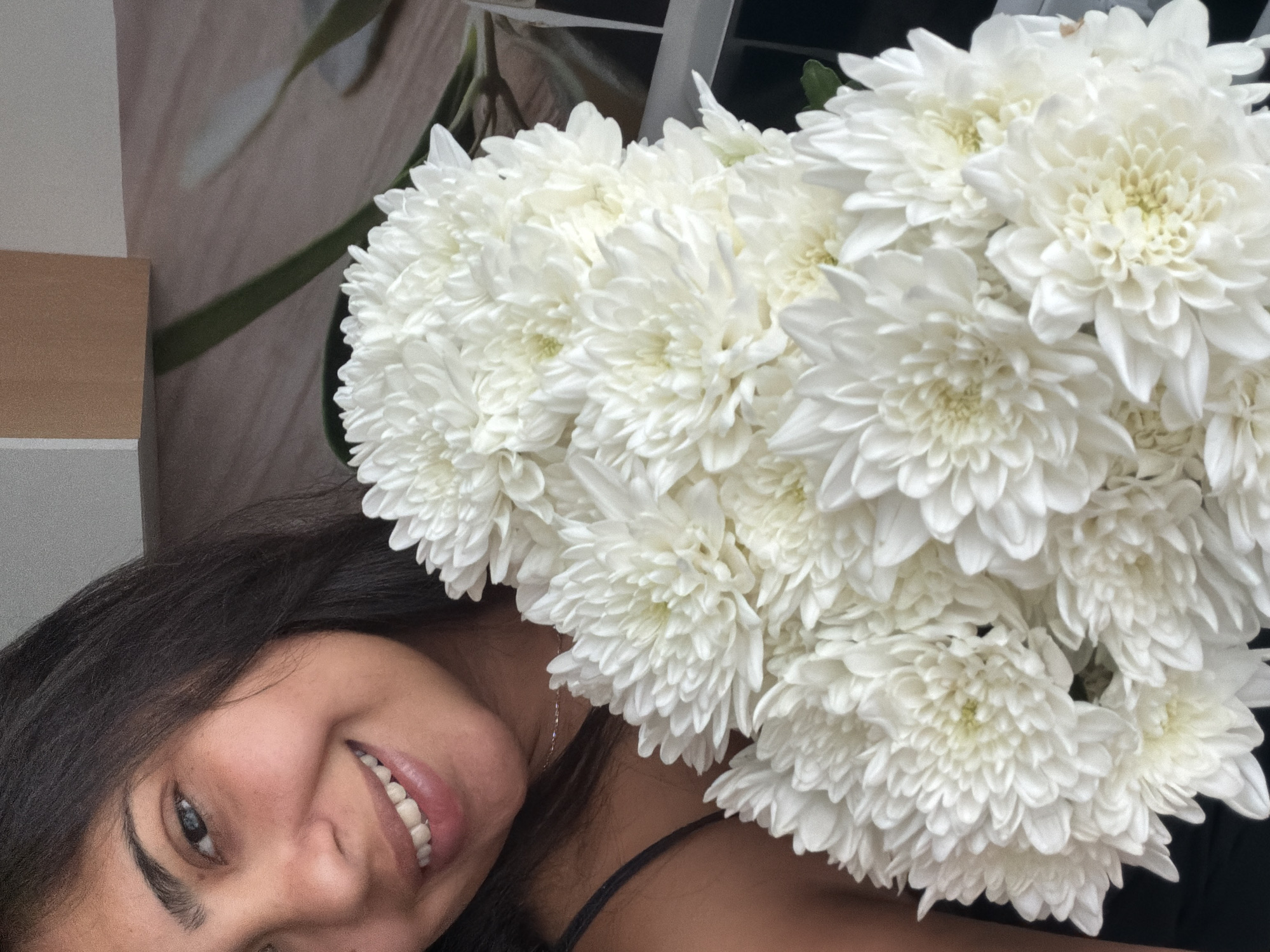 A close-up, slightly blurred, medium shot captures a young woman with dark hair and blue eyes smiling at the camera. She is holding a large bouquet of white chrysanthemums. Her head is turned to her left, and the flowers obscure the lower half of her face. The background is out of focus, showing a light-colored wall with a wooden-patterned surface and a hint of greenery. There is no indication of the city of Duisburg, Germany, or any specific urban environment.
