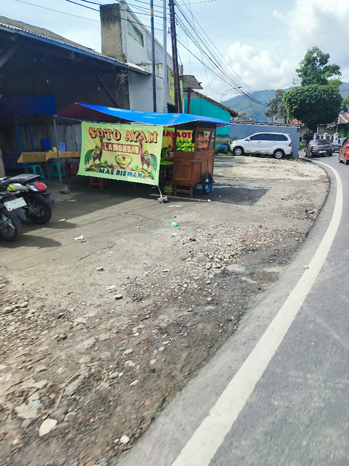 The image shows a street scene in Rancabali, Indonesia, located at the coordinates -7.122 latitude and 107.43 longitude. A food stall with a blue awning is set up on the side of the road, displaying a large yellow banner that reads "Soto Ayam Lamongan Mas Riswan". The stall is selling Soto Ayam, a popular Indonesian soup dish. A wooden cart with a canopy is situated next to the stall and appears to be part of it. A white car is parked on the opposite side of the street. The road is paved with asphalt and has a white line separating lanes. The surrounding environment is characterized by buildings, trees, and power lines. The ground in front of the stall is mostly gravel, with scattered debris.