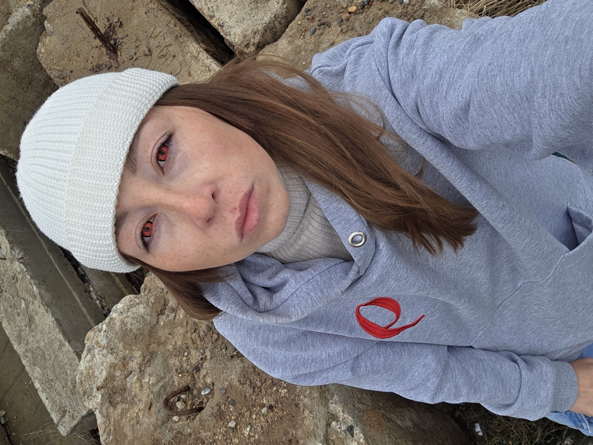 This tilted, medium close-up portrait features a young woman with reddish-brown hair, looking directly into the camera. She wears a cream-colored knitted beanie and a light gray hooded sweatshirt over a beige turtleneck or high-neck top. Her most striking feature is her vivid red eyes, contrasting with her otherwise natural facial appearance, which includes visible freckles on her nose and cheeks. Her lips are a soft pink. The gray hoodie has a distinct red embroidered logo on the left chest, resembling a stylized letter 'e' or a flowing ribbon. The woman's pose suggests she might be sitting or leaning back against the rough, weathered concrete blocks that form the background. These irregular blocks appear to be man-made, possibly industrial or construction debris, with some exhibiting rusty rebar. The overall lighting is soft and diffused, indicating an overcast outdoor setting. The image captures a blend of casual attire with a captivating, almost otherworldly, gaze.