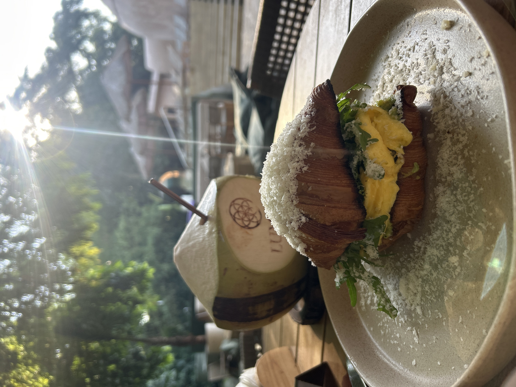 This image captures a close-up of a breakfast or brunch setting on a light brown wooden slatted table, located in the city of Unknown, Unknown. In the immediate foreground, a croissant sandwich rests on a round, light beige ceramic plate. The croissant is filled with fluffy yellow scrambled eggs, and green arugula leaves, with both the croissant and plate liberally garnished with grated white cheese. Behind the plate, a young, green coconut with a straw inserted stands upright on a dark coaster. A light-colored label on the coconut is partially visible, showing "LUM..." and a circular, geometric logo. The background is softly blurred by bokeh, revealing lush green foliage on the left and various outdoor structures on the right, including multiple thatched-roof umbrellas and light-colored buildings, indicative of a resort or outdoor cafe environment. Direct sunlight, originating from the upper right, casts a strong lens flare and bright highlights across the scene, illuminating the overall setting. No individuals are visible.