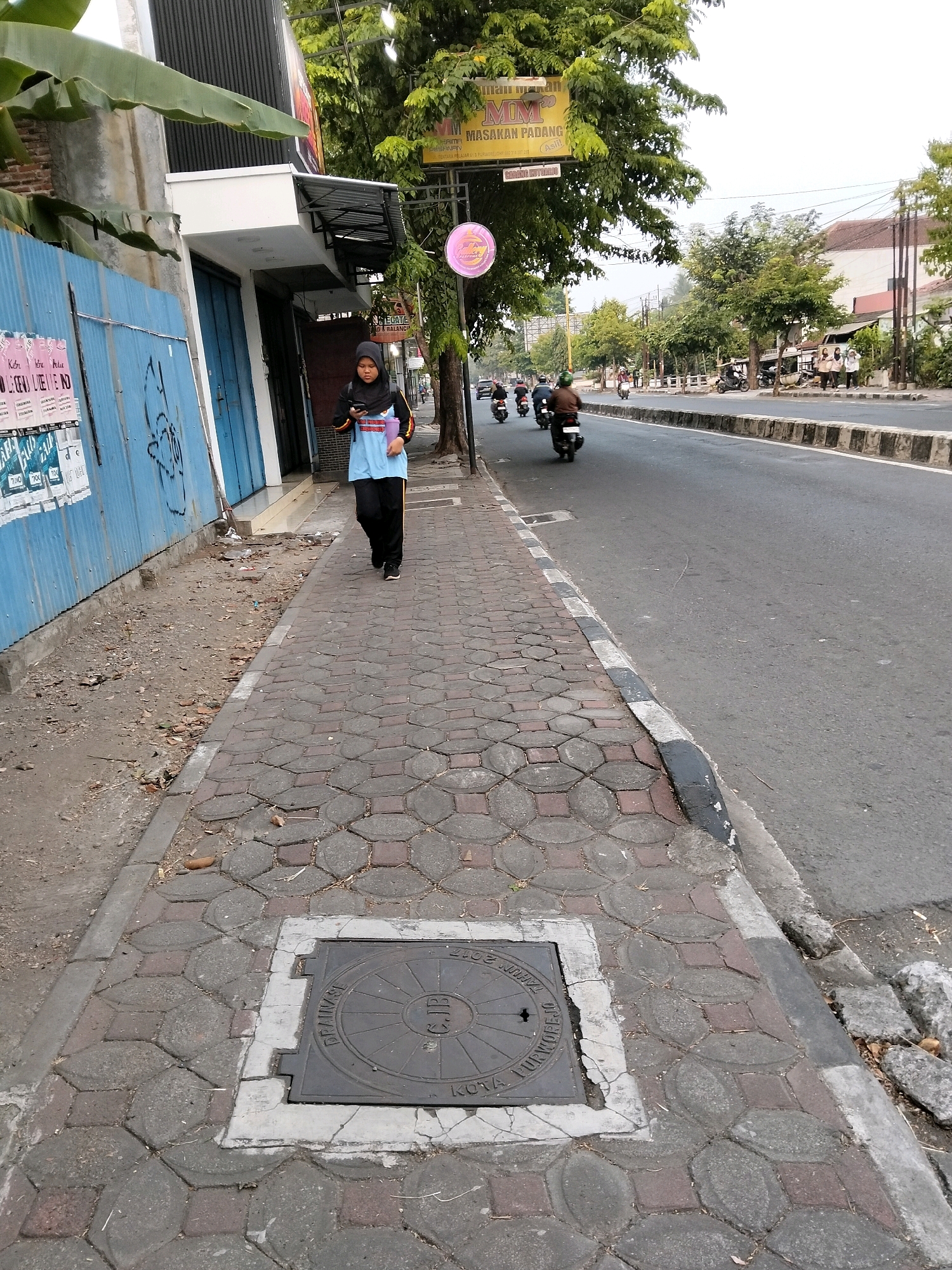 The image depicts a street scene in Purworejo, Indonesia. A young woman wearing a hijab, a light blue shirt, and black pants walks along a paved sidewalk. She is looking at her phone and holding a purple folder. The sidewalk is made of interlocking concrete pavers with a circular manhole cover embedded in it. To the left of the sidewalk is a blue corrugated metal wall with posters. In the background, a road is visible with several motorcycles and a few pedestrians on the opposite side. Trees line the street, and buildings can be seen in the distance. A yellow sign with Indonesian text, likely indicating a restaurant, is suspended from a pole. The overall scene suggests a typical day on a city street.