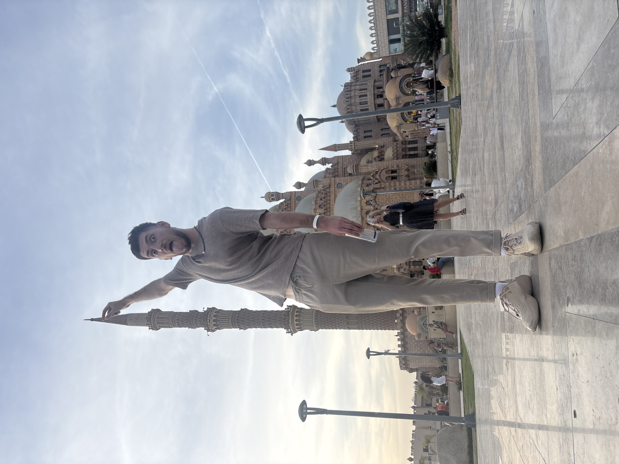 A man in a short sleeve brown t-shirt, light brown pants, and white sneakers is posed as if pinching the top of the building, which is in front of him. The building is located in Sharm el-Sheikh, Egypt. The background has other buildings that look similar in design to the first. The sky is light blue with some clouds. The floor is tile. Other people are walking around in the background.