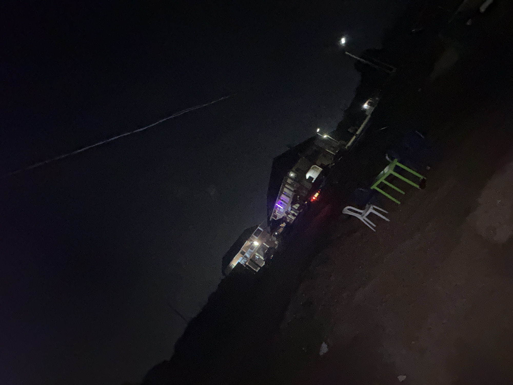 The image depicts a nighttime scene on a dirt road in Jos, Nigeria. Two multi-story buildings are visible, illuminated by exterior lights and some colored lights on the facade of the building on the right. The building on the left appears to have balconies. In the foreground, there are several plastic chairs and a table, suggesting an outdoor seating area. A street light is visible to the right of the frame. The sky is dark, with a faint linear streak, possibly a contrail, extending across it. The ground is uneven and appears to be unpaved.