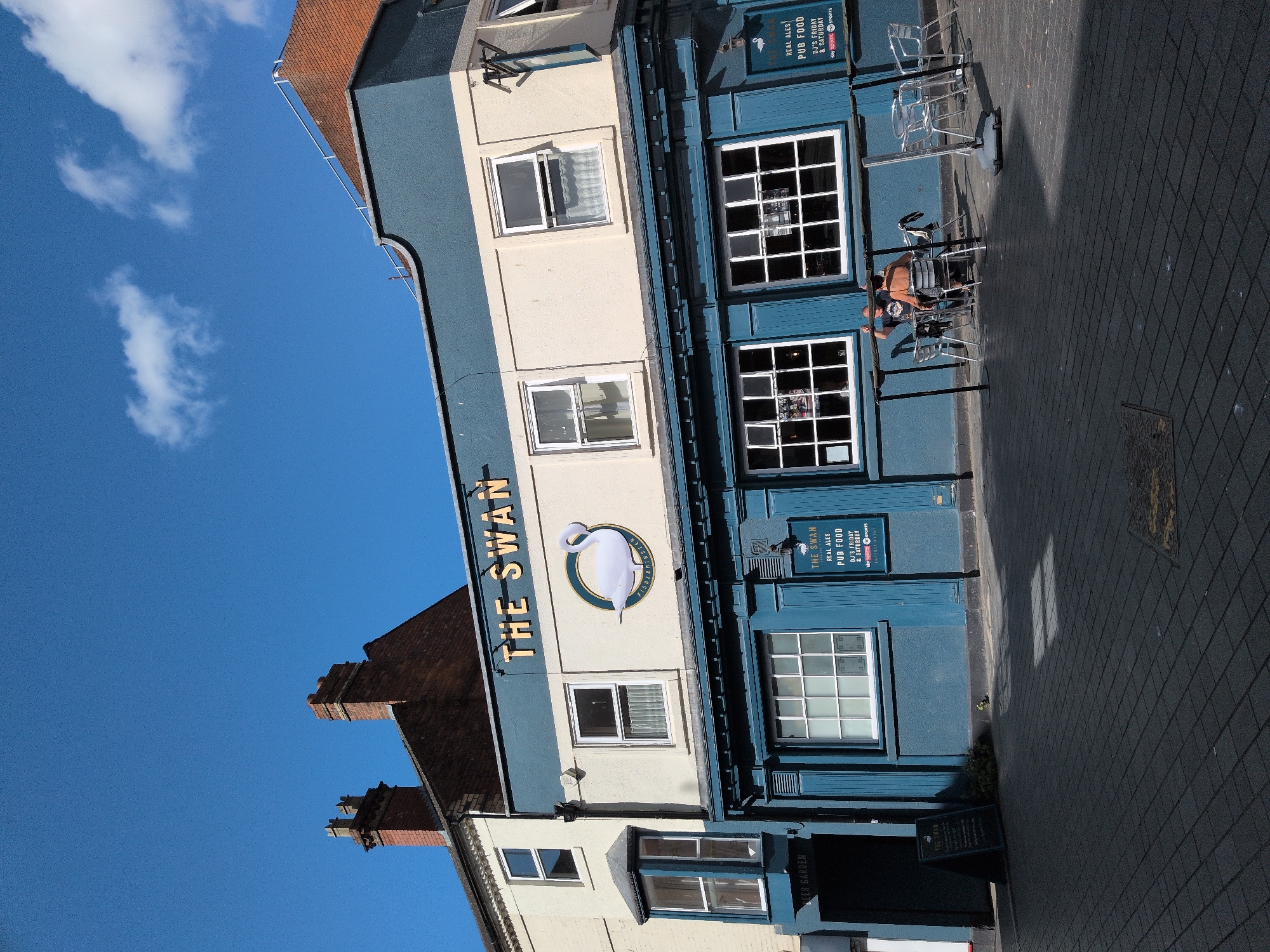 A pub named "The Swan" is depicted in a street scene. The pub has a blue and cream facade with "THE SWAN" written in large golden letters above the windows. A white swan logo is also visible on the building. Outside the pub, there are tables and chairs set up, and two people are seated at one of the tables. A sign with "BEER GARDEN" is visible to the left of the entrance. Another sign displays "THE SWAN REAL ALES PUB FOOD" with details about DJ nights. The pavement is paved with dark grey tiles, and there are shadows cast by the building and some objects. The sky is blue with a few white clouds. The location is Wyre Forest, United Kingdom.