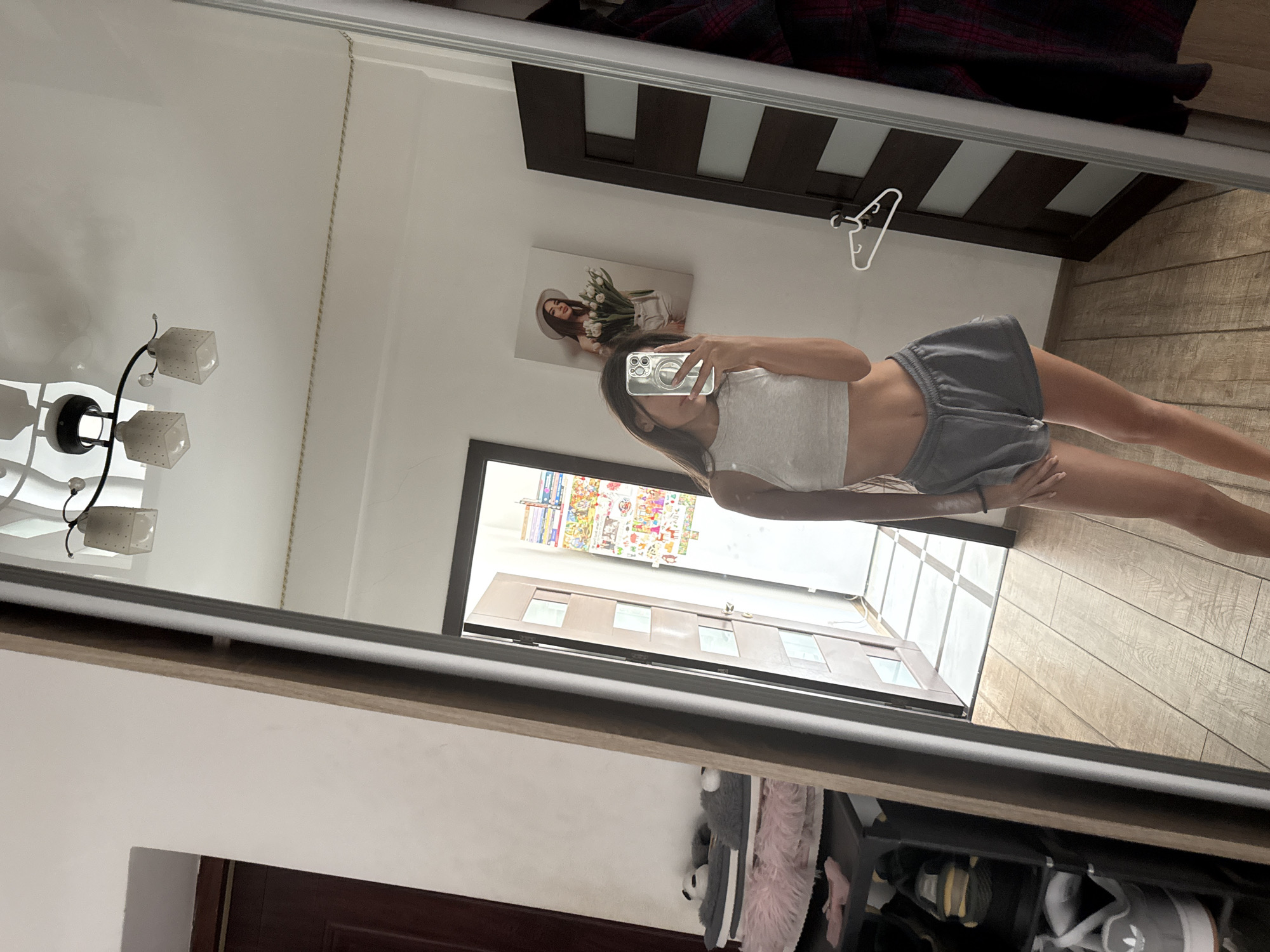 A young woman is taking a mirror selfie in a room. She is wearing a grey crop top and grey shorts. She is standing in front of a large mirror that reflects the room. The room appears to be a bedroom or a dressing area. There is a door behind her, and a shoe rack to her left. A painting of a woman holding flowers is on the wall behind her. The floor is made of wood planks. The overall lighting in the room is bright. This image was taken inside a residential property in Chisinau, Moldova.