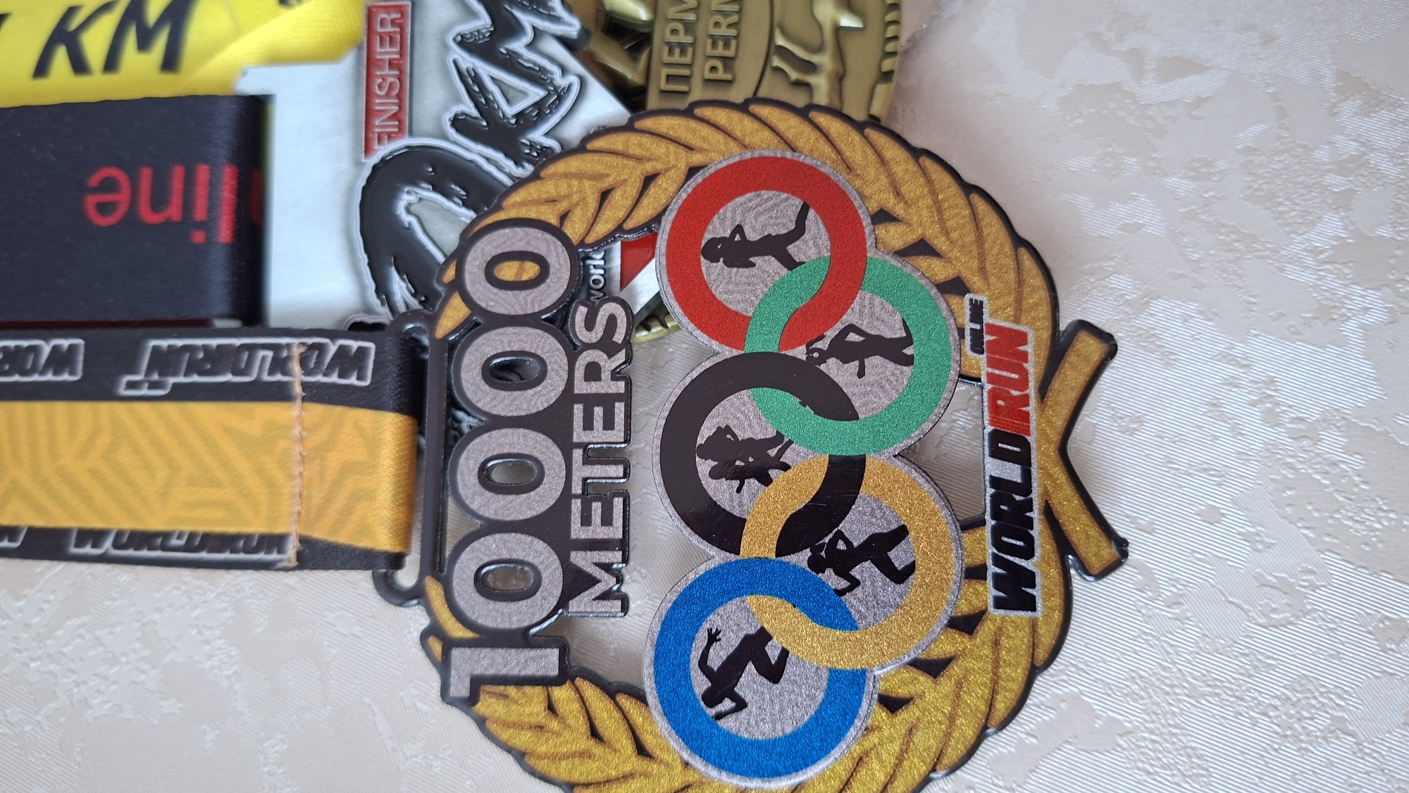 The image shows a close-up of several race medals. The most prominent medal is a "10000 METERS WORLDRUN" medal with a design resembling Olympic rings, each ring containing a silhouette of a runner. The rings are colored red, green, black, gold, and blue. This medal is surrounded by a gold laurel wreath. Other medals are partially visible in the background, including one with "FINISHER" and another with "ПЕРМЬ". The ribbon attached to one of the background medals is black with "online" in red. The ribbon attached to the 10000 meters medal is black and gold with "WORLDRUN" repeated. The medals are sitting on a textured surface. The "WORLDRUN" series takes place in Chaikovsky, Russia, according to other search results.