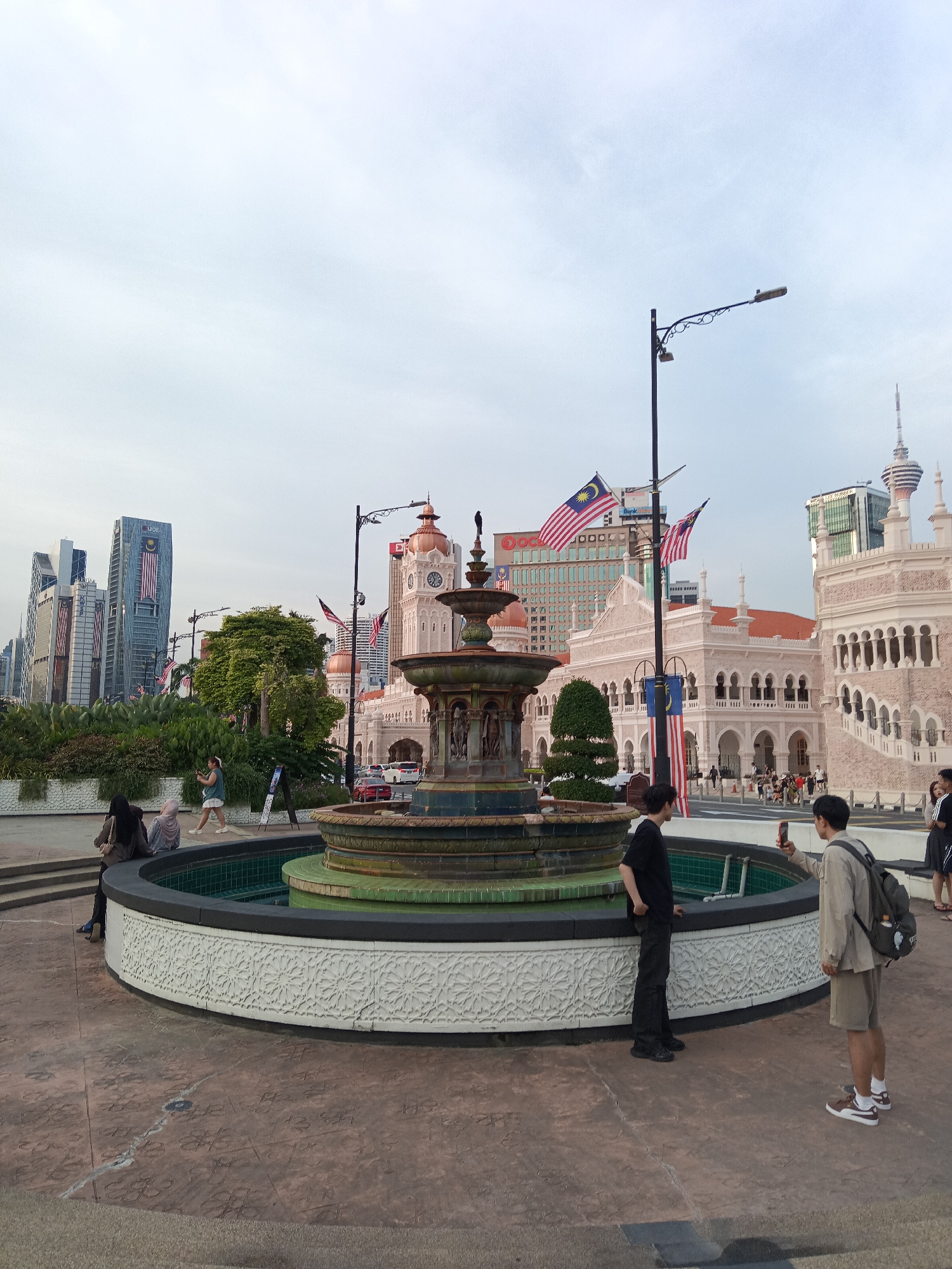 The image captures Merdeka Square in Kuala Lumpur, Malaysia, a historical landmark known for its colonial architecture and national significance. In the foreground, a circular fountain with multiple tiers is centrally located, its base adorned with intricate white tilework. The fountain's basin contains dark green water.
To the right of the fountain, two young men are standing. One, wearing a black t-shirt and dark pants, faces the fountain. The other, dressed in a beige jacket, shorts, and sneakers, holds up a smartphone and appears to be taking a picture or video of the scene. He is carrying a backpack.
In the middle ground, several individuals are seen near the fountain and walking further into the square. A woman in a light blue dress is seen walking away from the fountain, holding a phone. Further back, a group of women are seated near the edge of the fountain's decorative base.
The background is dominated by the Sultan Abdul Samad Building, a prominent colonial-era structure with distinctive arches and a clock tower. To the left of this building, a modern skyline rises, featuring several tall skyscrapers, some adorned with the Malaysian flag. Malaysian flags are also visible on poles throughout the square. Green trees and landscaping add to the urban park setting. The sky is overcast, suggesting a diffused light condition. The ground is paved with a reddish-brown material with a patterned texture.