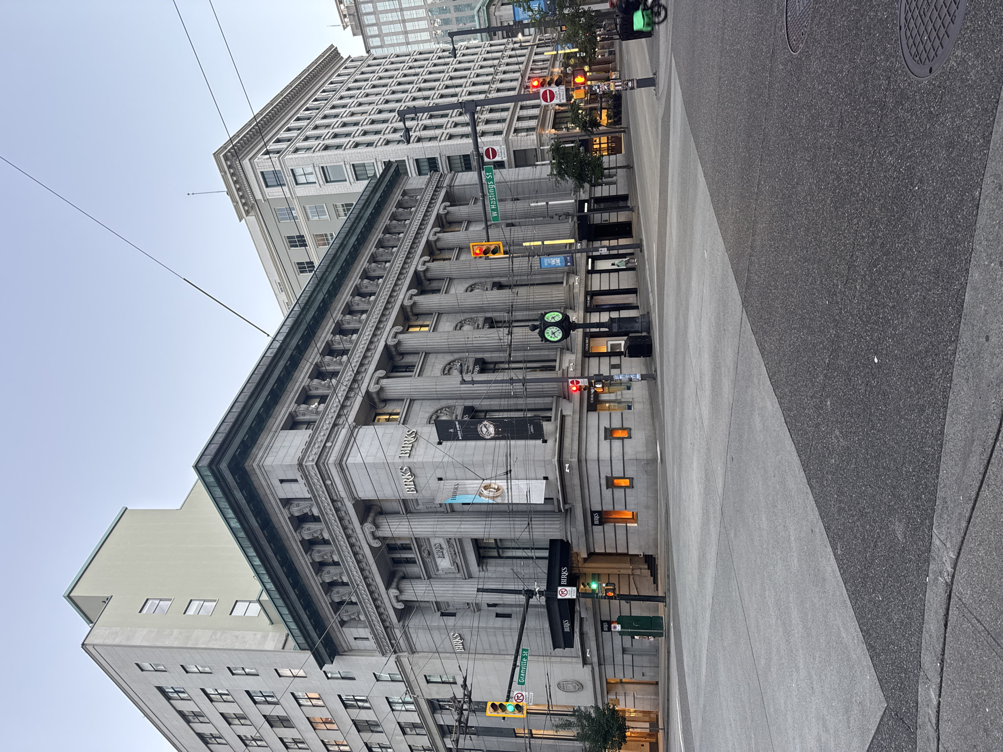 This is a medium-angle, eye-level, horizontal shot of a street intersection in Vancouver, Canada. The primary subject is the Birks building, an ornate neoclassical structure with prominent columns and decorative friezes. The building is on the corner of Granville Street and West Hastings Street, indicated by street signs visible in the image. The sky is clear and pale, suggesting either early morning or late evening.
On the left side of the image, a traffic light shows a green signal for eastbound traffic on Granville Street. A street clock with Roman numerals is positioned on the sidewalk near the Birks building. Several banners hang from the building, some featuring the Birks logo and advertising luxury watches.
Across the intersection, on the right side of the image, a traffic light shows a red signal for eastbound traffic on West Hastings Street. A person on a green bicycle is visible moving away from the camera at the far right of the image. There are other pedestrians in the distance, but they are not clearly discernible.
The sidewalks and street are paved with concrete and asphalt, with some contrasting patterns in the paving stones at the intersection. Utility poles with overhead wires are present throughout the scene, indicating an urban environment with active electrical infrastructure. The overall impression is of a quiet, early-morning or late-evening urban setting in a commercial district.