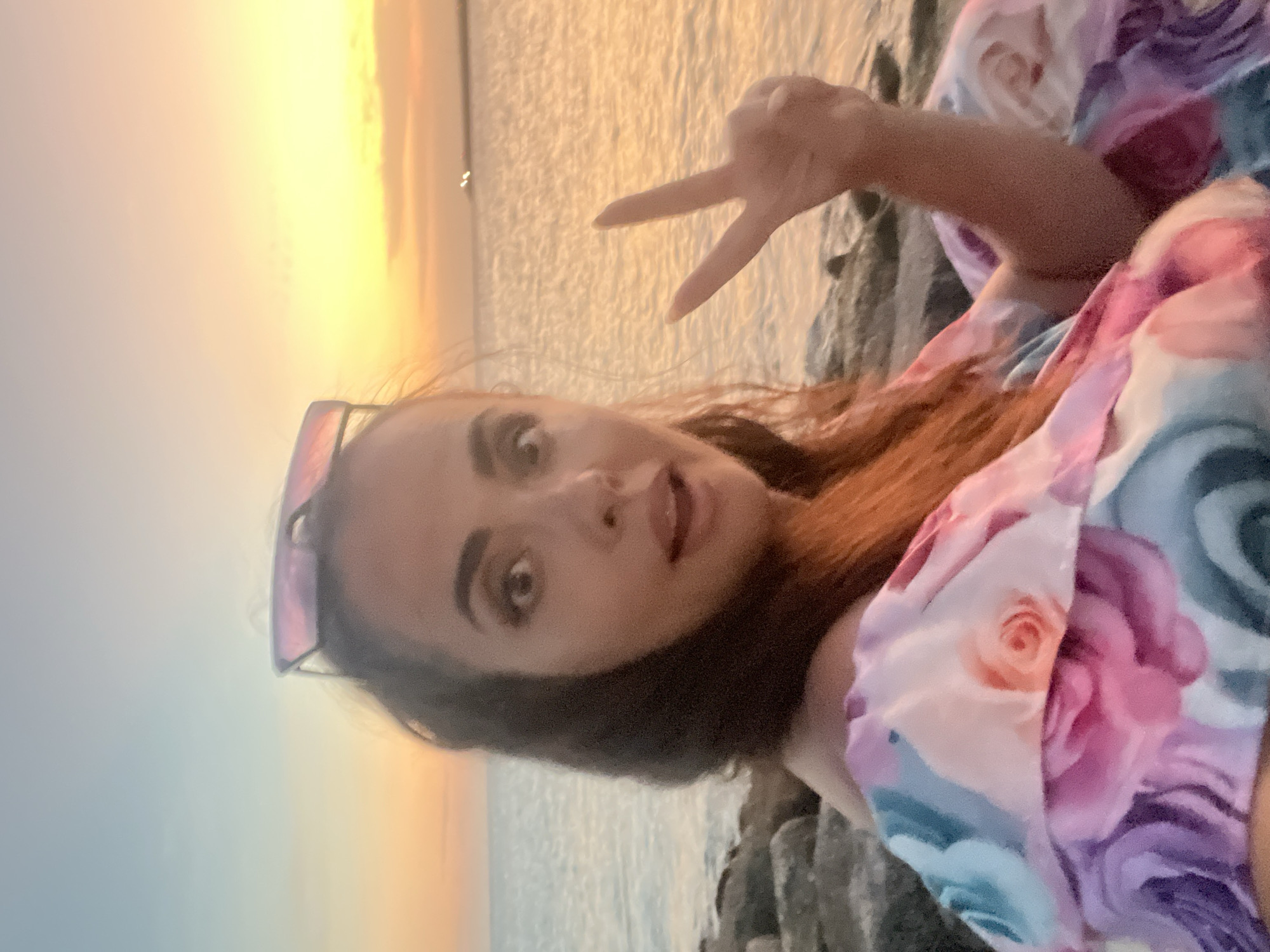 A medium shot captures a woman with auburn hair and dark eyes looking directly at the camera with an open mouth, her right hand raised in a peace sign. She is wearing a floral off-the-shoulder dress with pink, purple, and blue roses. Her sunglasses are perched on top of her head. The woman is seated on rocks near the water's edge, with the ocean and a warm, orange-yellow sunset in the background. The location is likely Kuta, Indonesia, based on the warm coastal atmosphere and sunset common to the region.