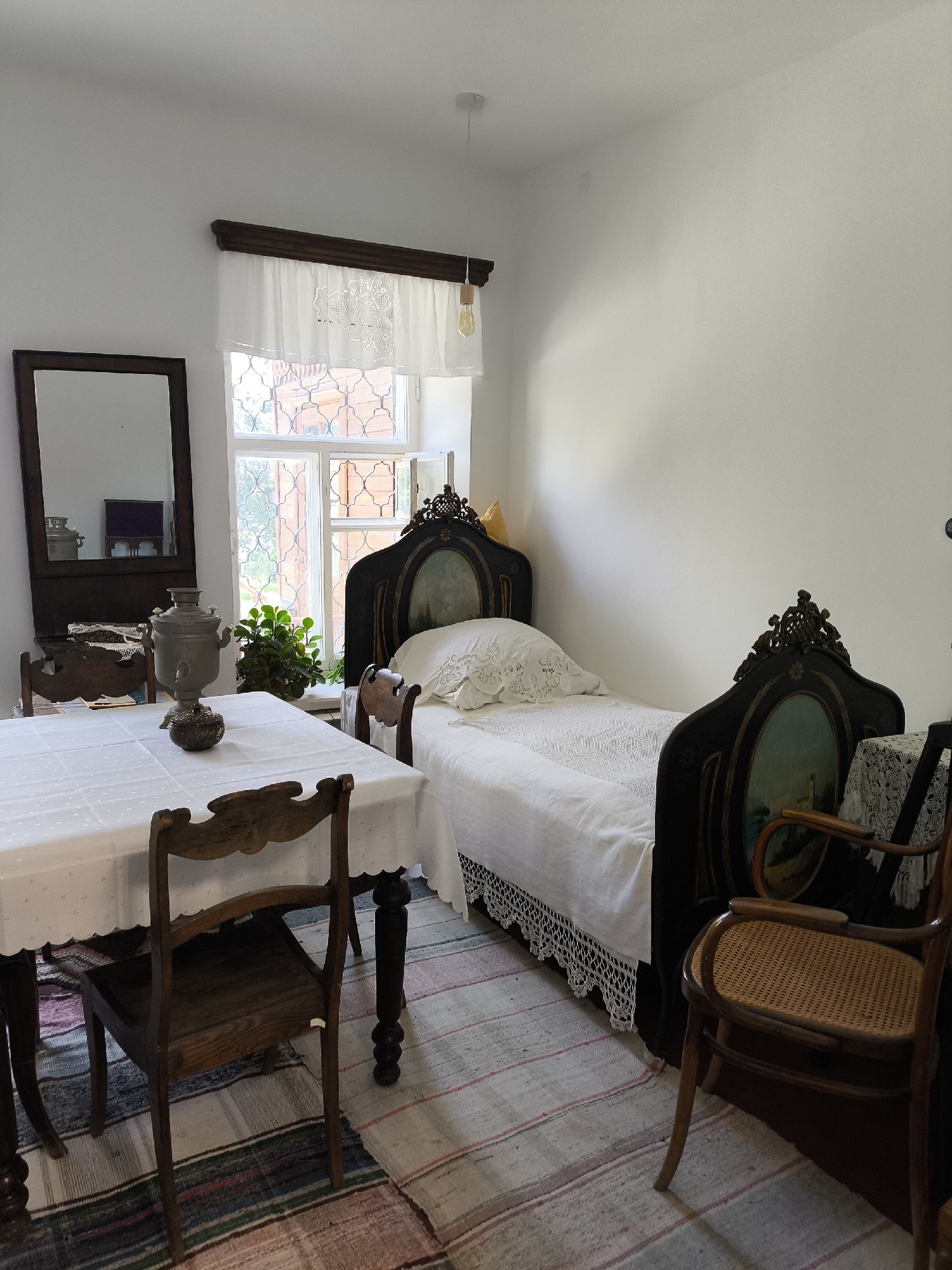 This is a photograph of a room that appears to be a historical exhibit in Yekaterinburg, Russia. The room is furnished in a traditional style, likely representing a dwelling from the late 19th or early 20th century.
In the foreground, a wooden dining table is covered with a white tablecloth that has a patterned border. Two wooden chairs are placed at the table; one is partially visible in the lower left, and another, with a carved wooden back, is on the left side of the image. On the table sits a large, ornate silver samovar and a smaller decorative item.
To the right of the table, there is a narrow bed with a dark, intricately carved headboard and footboard, each featuring a painted oval landscape. The bed is dressed in white linens, including a white lace-trimmed bedspread and a white pillow with lace trim. Next to the bed, a wooden chair with a woven cane seat is positioned.
A large mirror in a dark wooden frame hangs on the wall to the left of the window, reflecting a part of the room, including what appears to be a cabinet or dresser. The window itself is multi-paned with decorative white lace curtains and a dark wooden valance. Outside the window, glimpses of greenery and an orange, latticed structure can be seen.
The floor is covered with a colorful striped rug. The walls are painted white, and a light fixture with an exposed bulb hangs from the ceiling. The overall impression is one of a preserved domestic interior, suggesting a historical museum or a preserved historic house. The image captures a static scene, with no people present.