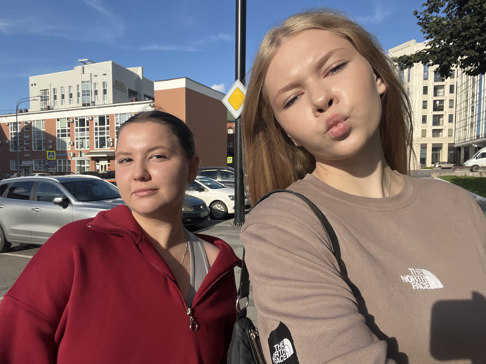 Two young women are taking a selfie outdoors in a city environment. The woman on the left is wearing a red hooded sweatshirt and a grey sports bra. She has dark hair pulled back in a bun and is looking at the camera with a neutral expression. The woman on the right has blonde hair and is wearing a light brown sweatshirt with a "The North Face" logo on the chest. She is puckering her lips and looking at the camera. In the background, there are modern multi-story buildings, a street sign, a traffic pole, parked cars, and trees. The sky is blue and clear, indicating a sunny day. The image appears to have been taken on a street in Perm, Russia, based on the street signs visible in the background which are in Russian.