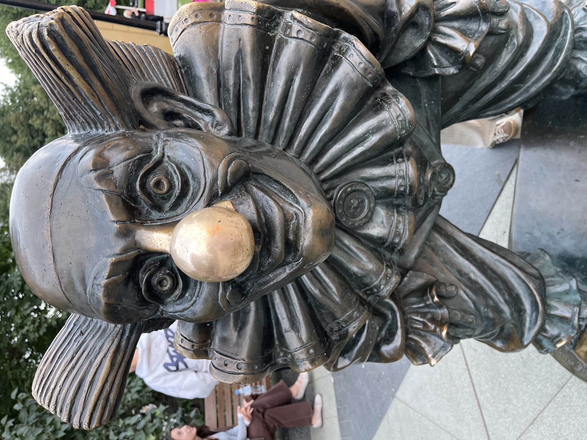 This is a close-up, low-angle, outdoor photograph of a bronze sculpture of a clown in a park in Moscow, Russia. The sculpture is detailed, depicting the clown with exaggerated features, including a large, round, metallic nose, wide eyes, a broad grin, and a ruffled collar. The clown's hair is stylized with prominent ridges.
In the background, two individuals are visible. To the left, a young woman with dark hair is sitting on a park bench, wearing a light blue top and brown pants, with her hands clasped in her lap. To her right, a person in a white hoodie and dark pants stands behind the bench. The environment appears to be a public park, with trees and foliage visible behind the people. The ground in the foreground is paved with a patterned surface of gray and dark gray squares. The overall lighting suggests it is daytime, possibly overcast.