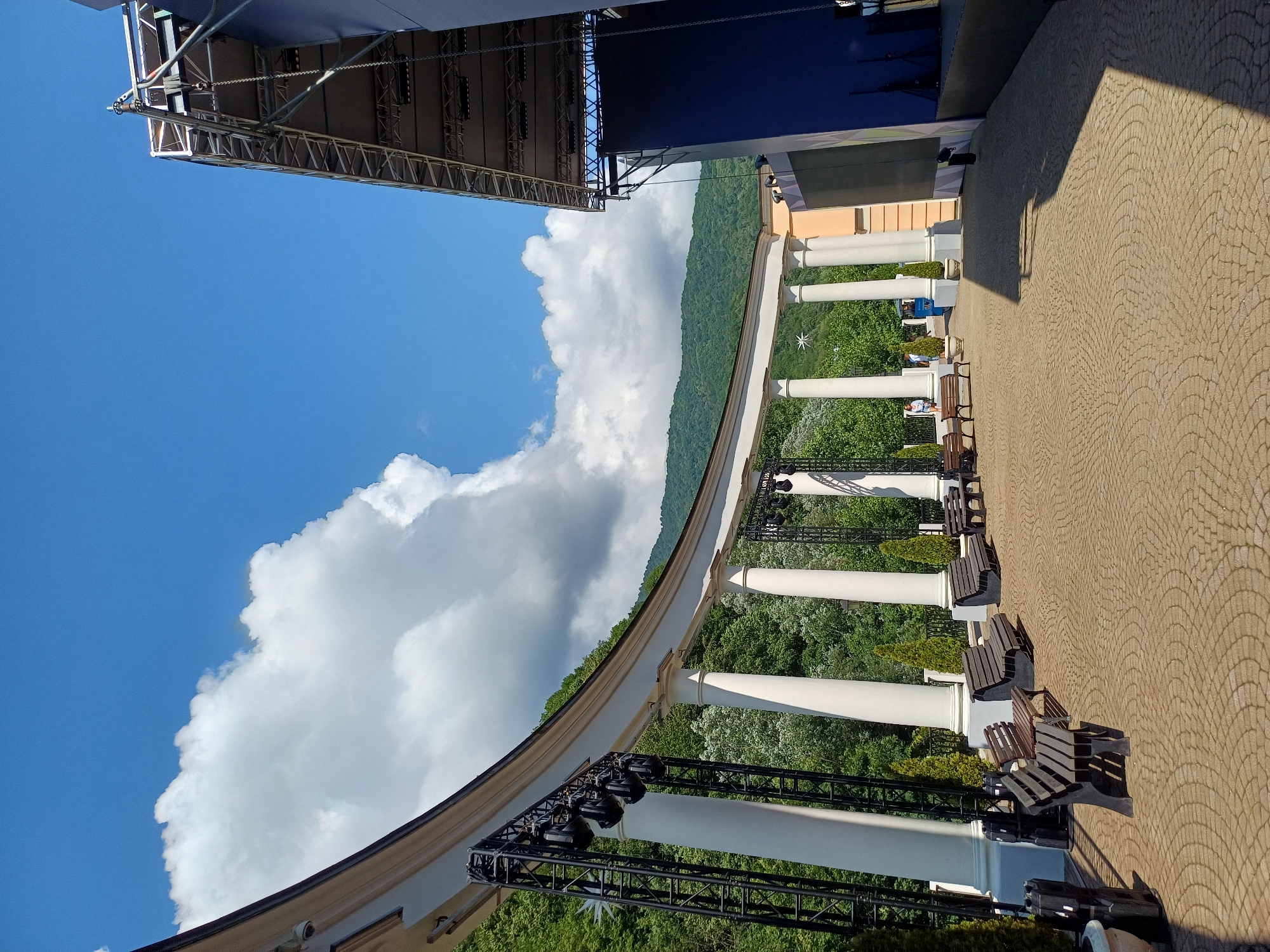 This is a low-angle, full shot of an outdoor amphitheater area in Estosadok, Russia, on a sunny day with scattered clouds. The curved colonnade, composed of white pillars supporting an arch, dominates the left side of the frame. On the colonnade, lighting rigs with multiple spotlights are mounted. Beyond the colonnade, a dense, green forest covers rolling hills, leading up to a mountain range in the background under a bright blue sky with prominent white clouds.
In the foreground, a paved area with a circular pattern is visible, with several dark wooden benches arranged on it. To the right of the colonnade, a large, blue stage structure with a metal roof and truss system is present, suggesting preparation for an event. A few small, conical evergreen trees are planted in decorative pots. Two individuals are visible in the distance, one standing and another walking. The overall scene suggests a public outdoor venue designed for performances or gatherings, set against a natural, mountainous backdrop.