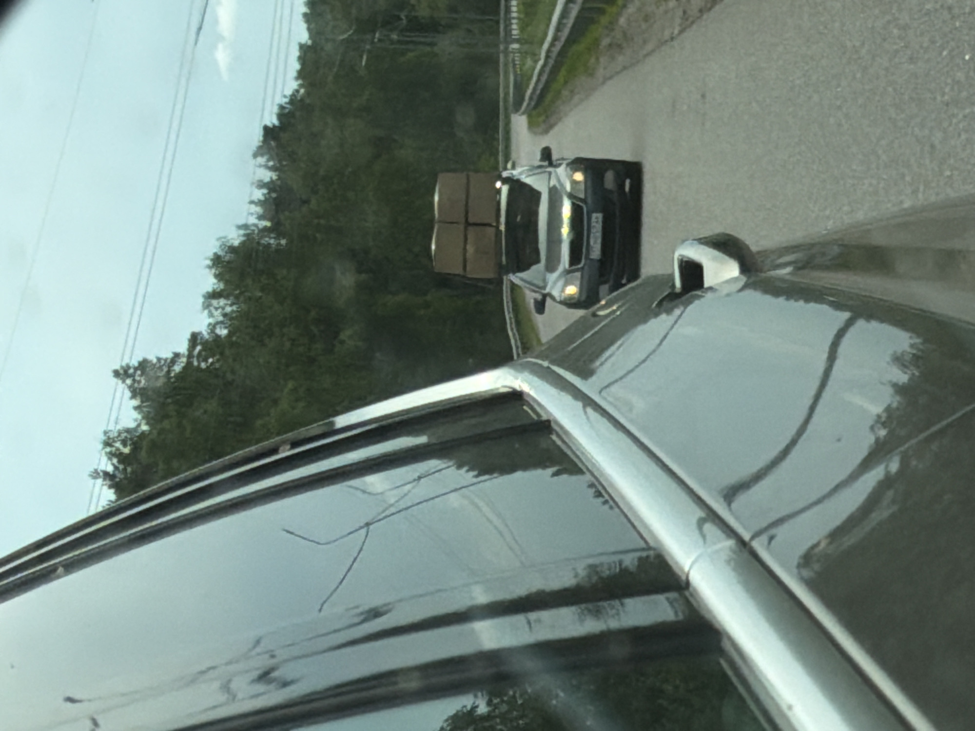 The image shows a car driving on a paved road. Another car is visible ahead, carrying several cardboard boxes on its roof. The background features a forest area and some power lines. The photo was taken from inside a car. There is no evidence to suggest the location is Plastik, Russia.