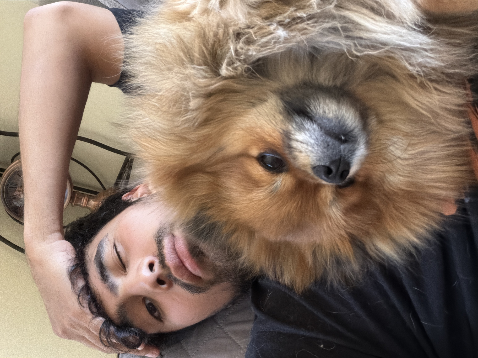 A man lies on his back, partially on a grey quilted surface, wearing a black shirt. He winks with his right eye. Above him is a dog with brown and tan fur. An analog clock sits on a table behind the man's arm. The specific location of the image in Cuautla, Mexico is indiscernible.