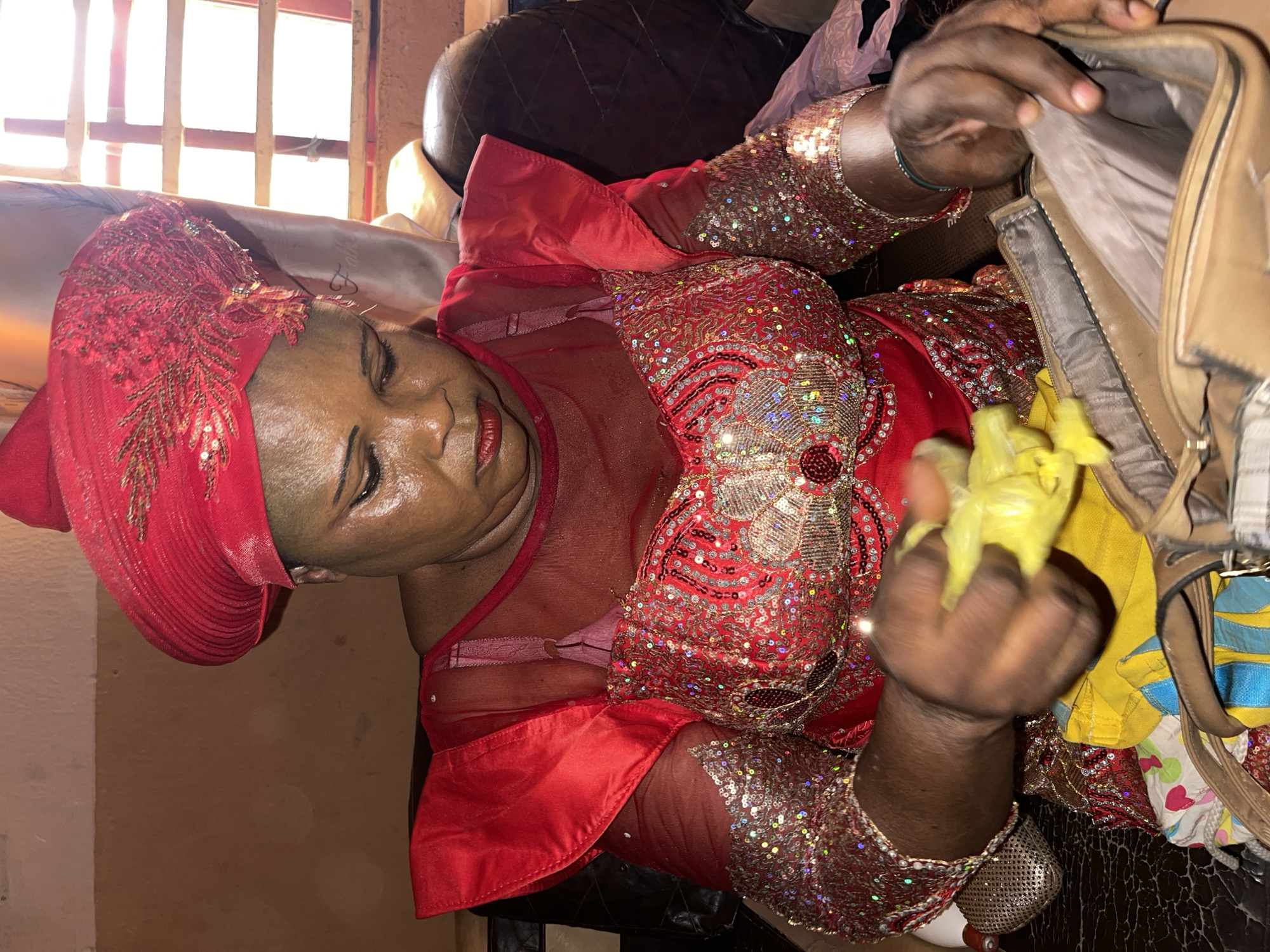 A middle-aged woman in Muchiya, Nigeria, is seen in an indoor setting. She is dressed in a red, heavily sequined, semi-transparent dress and matching headwrap. She is looking inside a brown handbag and holding a crumpled yellow object. A window with a barred frame is visible in the background.