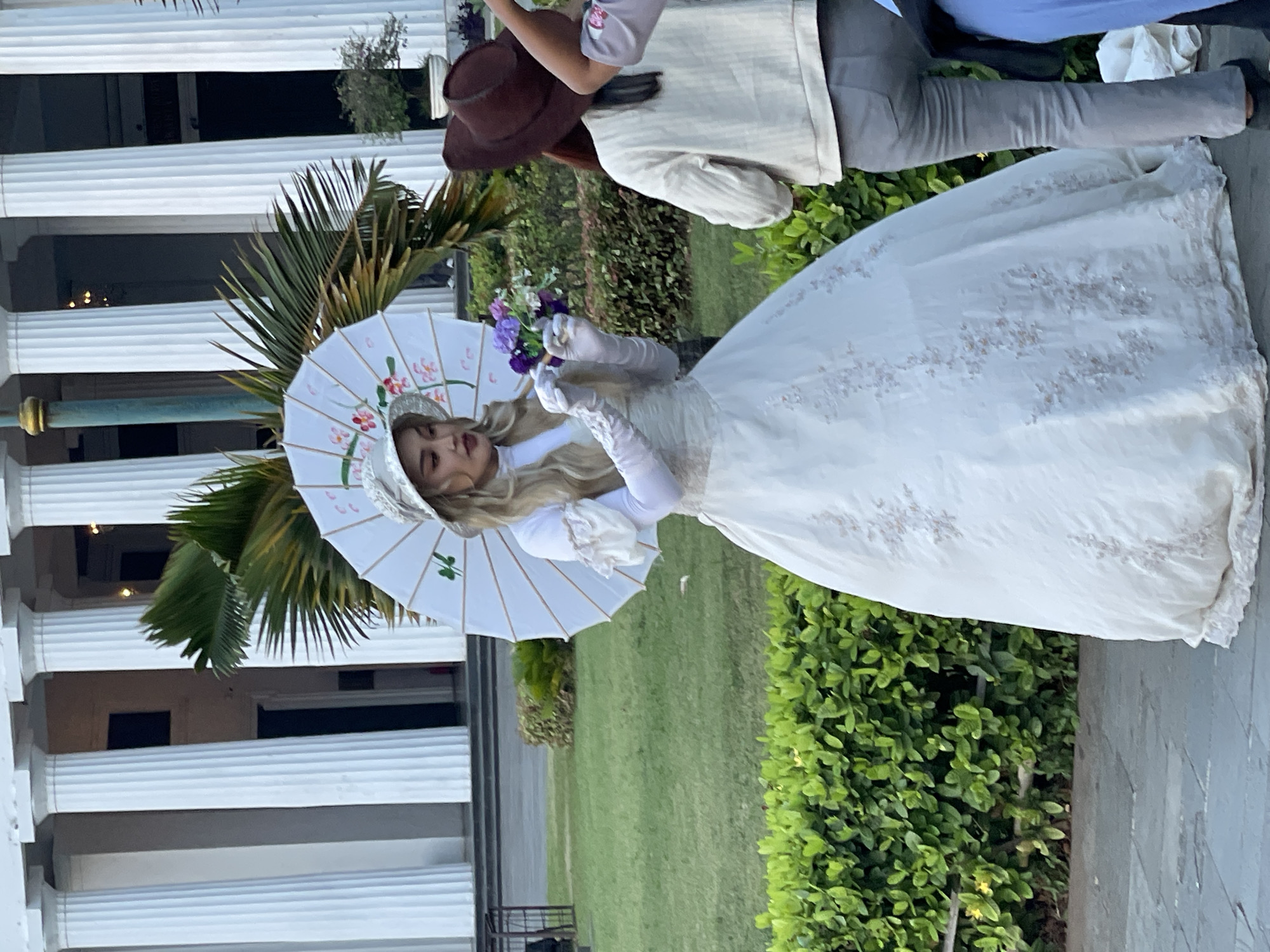 In West Jakarta, Indonesia, in front of the Fatahillah Museum, a woman in a white vintage-style dress and a hat stands holding a white parasol decorated with floral patterns. She wears white gloves and holds a bouquet of purple flowers. To her right, another person in a cream-colored jacket and brown hat is visible, and partially to the right of that person, another individual in a blue garment is also present. The background features a building with large white columns, lush green bushes, and a palm tree. The ground is paved with stone tiles.