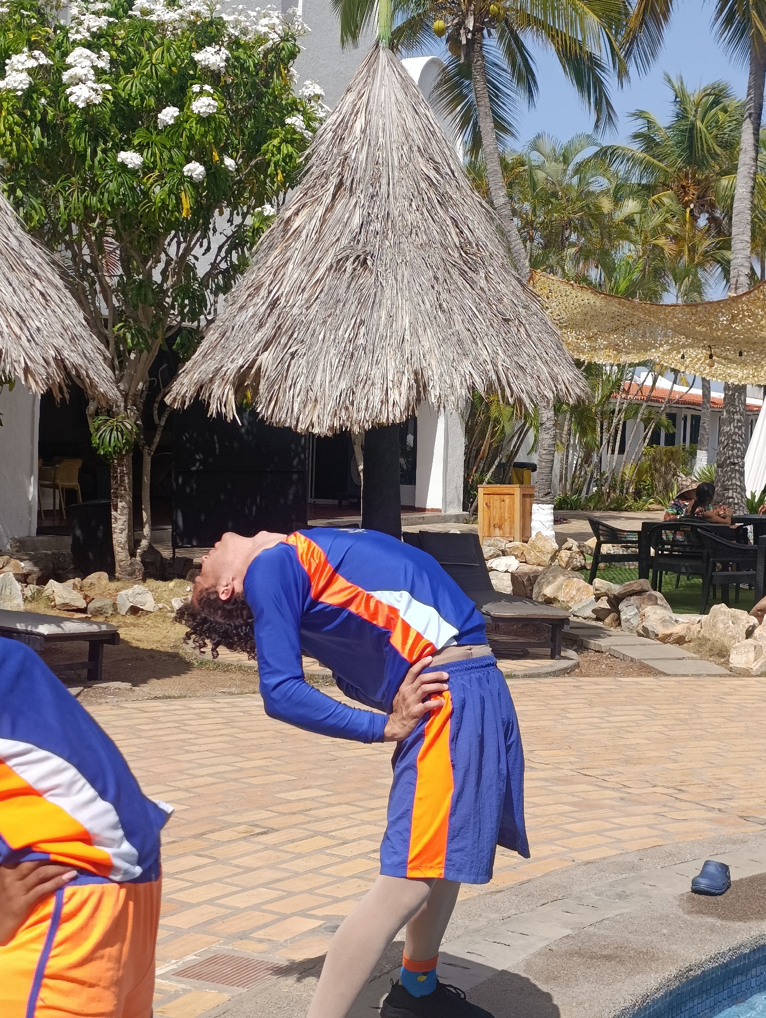 The image shows a person, likely male, wearing a blue and orange outfit, bending backward. The scene appears to be outdoors, in a resort setting. There are thatch-roofed structures nearby, and palm trees are visible in the background. The person is on a brick-paved area. The location could be in Porlamar, Venezuela, based on the tropical landscape. Other people are seated in the background near the resort buildings.