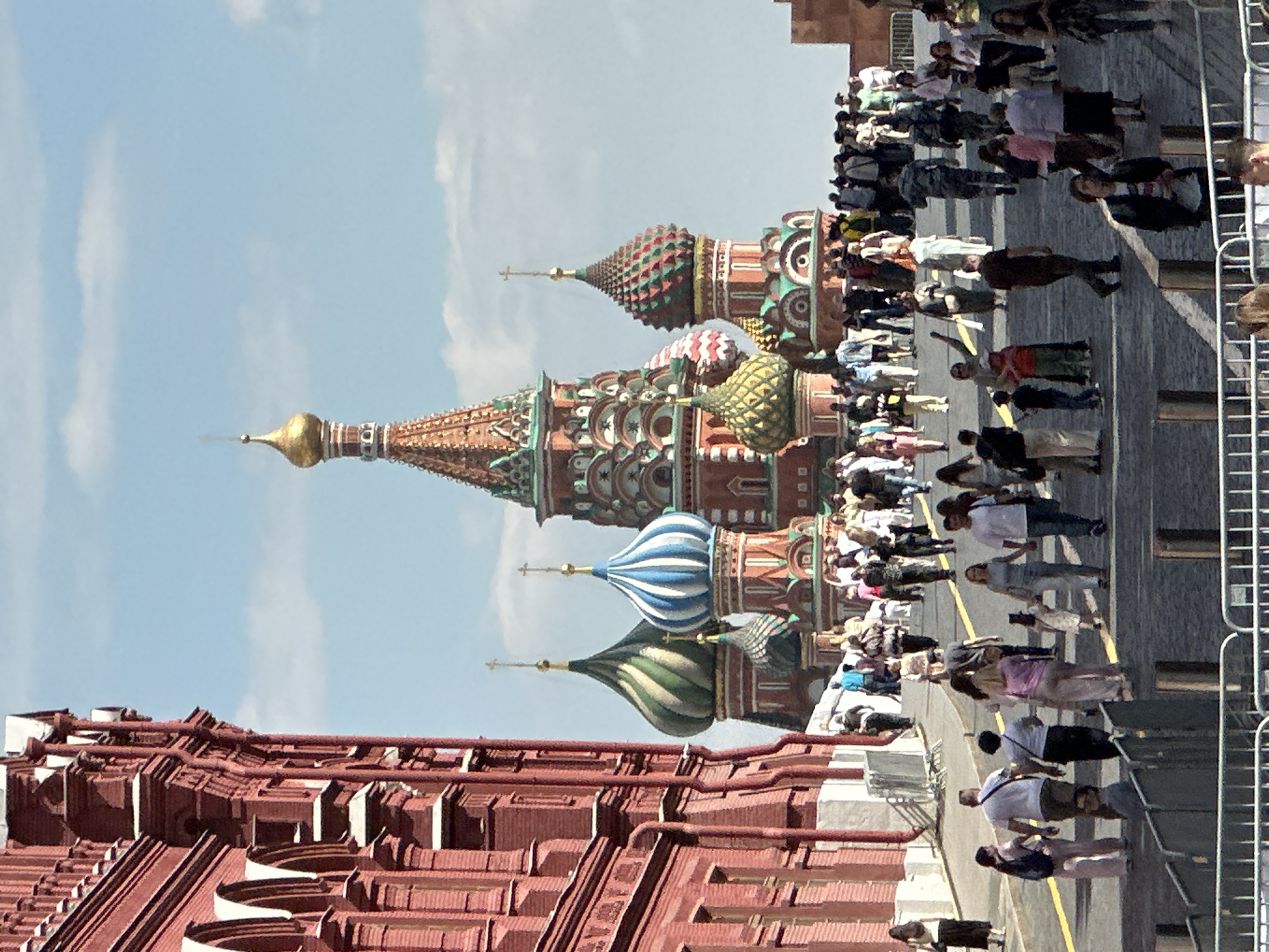 This daytime image captures a bustling scene in Moscow, Russia, featuring the iconic St. Basil's Cathedral and a large crowd. The cathedral, located in the background, showcases its vibrant and distinct onion domes and tented roofs, adorned with intricate patterns in shades of red, green, gold, blue, and white. Golden spires rise from several of the domes, set against a clear blue sky with sparse white clouds.
In the foreground and midground, numerous people, mostly adults dressed in casual clothing, are walking across a paved area, likely Red Square. Their movement suggests a typical day of sightseeing and tourism. To the left, part of a grand red brick building with ornate architectural details and a complex roof structure is visible, consistent with the historical buildings surrounding Red Square, such as the State Historical Museum or Kremlin walls. Metal railings line the right side of the scene, separating the walking crowd from another area. The bright sunlight indicates a clear and sunny day. No visible text is discernible in the image.