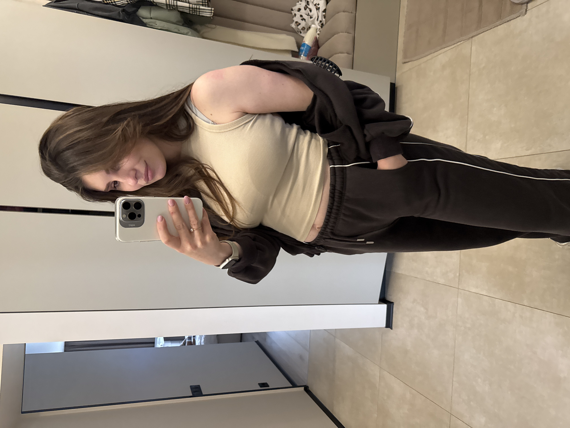 A young woman with long, dark brown hair is captured in a mirror selfie, taken indoors. She stands with her head slightly tilted, offering a gentle smile towards the camera of the light-colored smartphone she holds in her left hand. On her left wrist, she wears a light-colored smartwatch, and a ring is visible on her right ring finger.
She is dressed casually in a light beige or off-white ribbed tank top, layered under an open, dark brown or black zip-up hoodie. Matching dark brown or black sweatpants with a distinct white stripe down the side complete her outfit, with one hand casually placed in a pocket.
The setting appears to be a modern room, possibly a bedroom or dressing area, characterized by clean, light-colored (likely white or off-white) walls or furniture surfaces and light-colored tiled flooring. A dark horizontal line suggests a cabinet or closet design. In the reflection behind her, hints of other room elements are visible, including patterned textiles (one resembling leopard print) and a plaid item, indicating a comfortable and personal space. The word "TOY" appears backward as "ЯОТ" on the phone's casing in the reflection. The overall atmosphere is relaxed and casual.