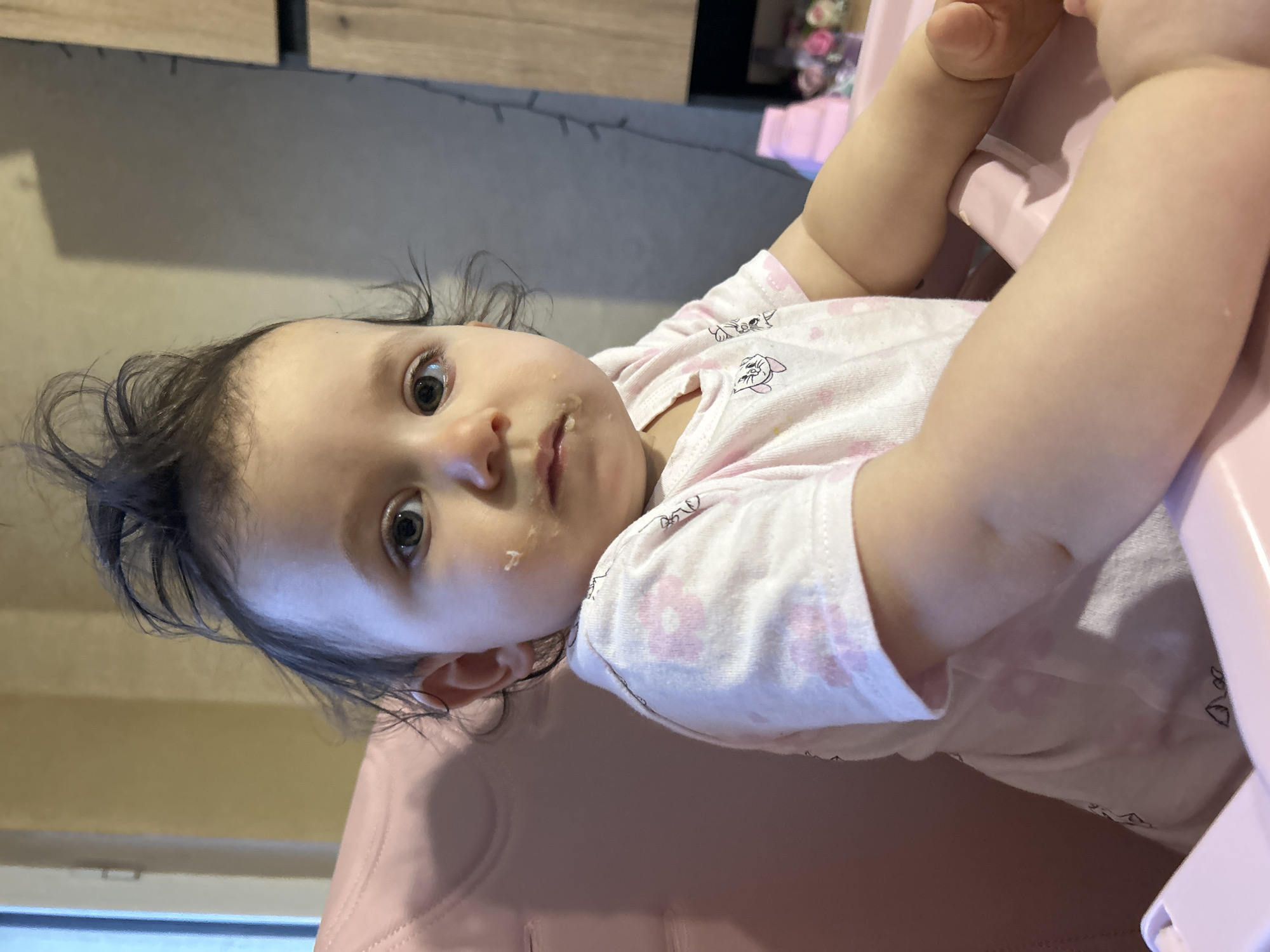 This image, taken in Sofia, Bulgaria, captures a baby girl seated in a pink high chair indoors. The baby, appearing to be under two years old, has dark, slightly disheveled hair and dark, observant eyes, looking directly at the viewer. She is wearing a light pink, short-sleeved onesie or top patterned with small white and pink designs, including what appear to be cartoon cats and possibly flowers. There is a small amount of white food residue on her face around her mouth, suggesting she has recently eaten or is currently eating.
The high chair is a pastel pink, providing a soft color contrast to the baby's attire. In the background, sections of a wooden cabinet or shelf are visible, alongside a blurred area that contains decorative elements, possibly flowers, on another surface. The lighting within the room appears ambient, suggesting it is daytime or early evening. The scene depicts a quiet domestic moment, likely in a kitchen or dining area. No other individuals or visible text are present in the image. The baby's expression is neutral and focused on the camera.
