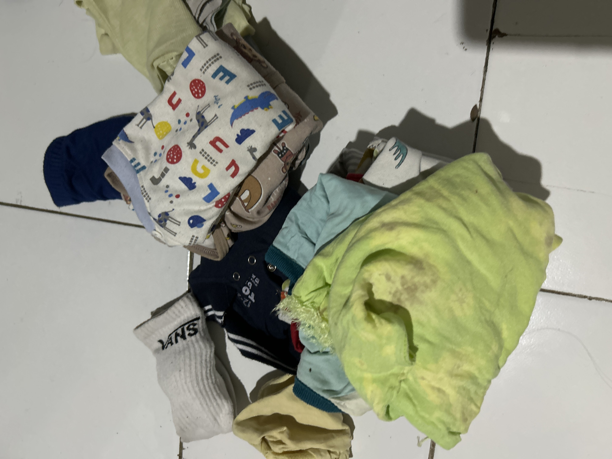A pile of laundry is scattered on a white tiled floor in Depok, Indonesia. The laundry includes a white sock with "VANS" written on it, a dark blue sock, a light blue and white patterned garment with animal prints, a dark blue shirt with snaps, a light yellow shirt, and a large, crumpled light green garment with brown stains. Some of the garments are folded, while others are bunched up. The lighting suggests an indoor setting, possibly a home.