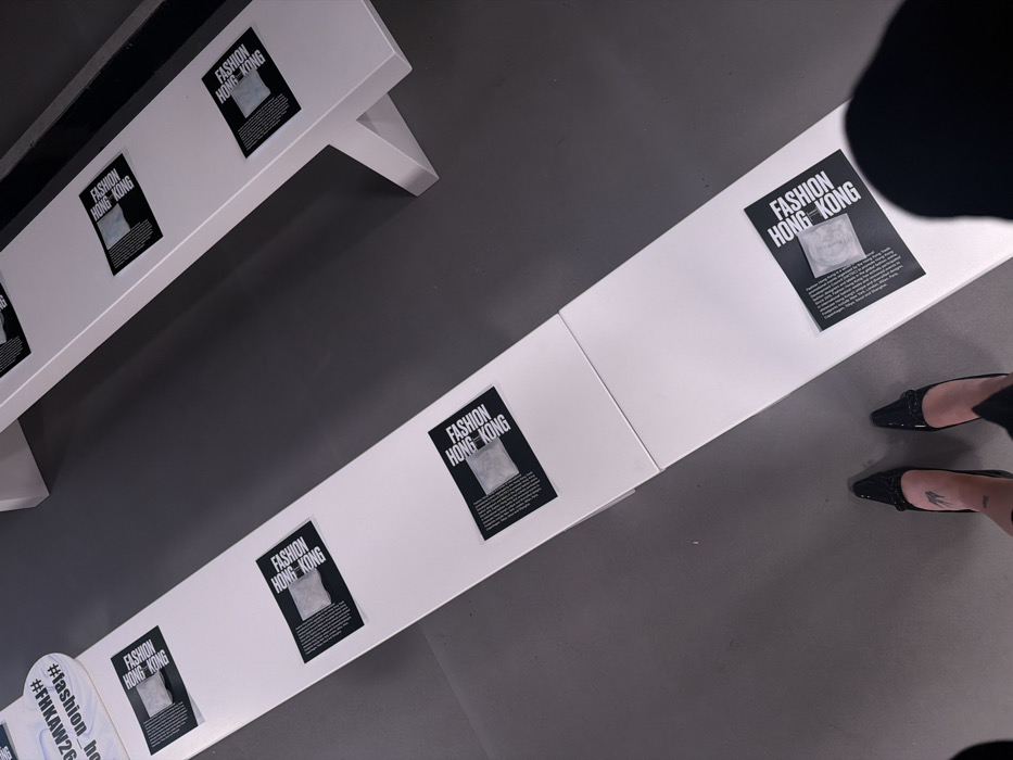 An overhead shot captures a minimalist indoor setting, likely a gallery, exhibition space, or event. The primary elements are two long, white, rectangular display structures, resembling benches or low tables, set against a dark gray floor.
The closer display structure extends diagonally from the mid-left to the right side of the frame, segmented into several sections. The further display structure is positioned behind and to the left of the closer one, also diagonally oriented.
Affixed to the top surface of both white structures are multiple black and white posters. Each poster features the prominent white text "FASHION HONG KONG" in a bold, sans-serif font, against a black background. Below this text, a square, light gray/white object, appearing to be crumpled fabric or a textured material, is centered. Beneath this image, a block of small, unreadable text is present on each poster. There are at least six such posters visible on the closer structure, and at least two on the further one.
On the far left, at the start of the closer white structure, a rounded white sign is visible. It displays hashtags in black text, including "#FHKAW26" and "#fashion_ho" (likely part of "#fashion_hongkong" or similar).
In the bottom right corner of the image, a person's feet are visible, standing on the dark gray floor next to the closest display structure. The person is wearing black, pointed-toe high-heeled shoes with a subtle embellishment on the vamp. A small, dark tattoo, possibly a stylized fish or similar design, is visible on the top of the right foot/ankle.
The lighting appears even, suggesting an indoor setting with artificial illumination, casting soft shadows beneath the white structures. The overall color palette is restricted to black, white, and dark gray, contributing to a clean and modern aesthetic. The arrangement of the posters and the presence of the hashtags suggest an event or exhibition focused on fashion from Hong Kong.
