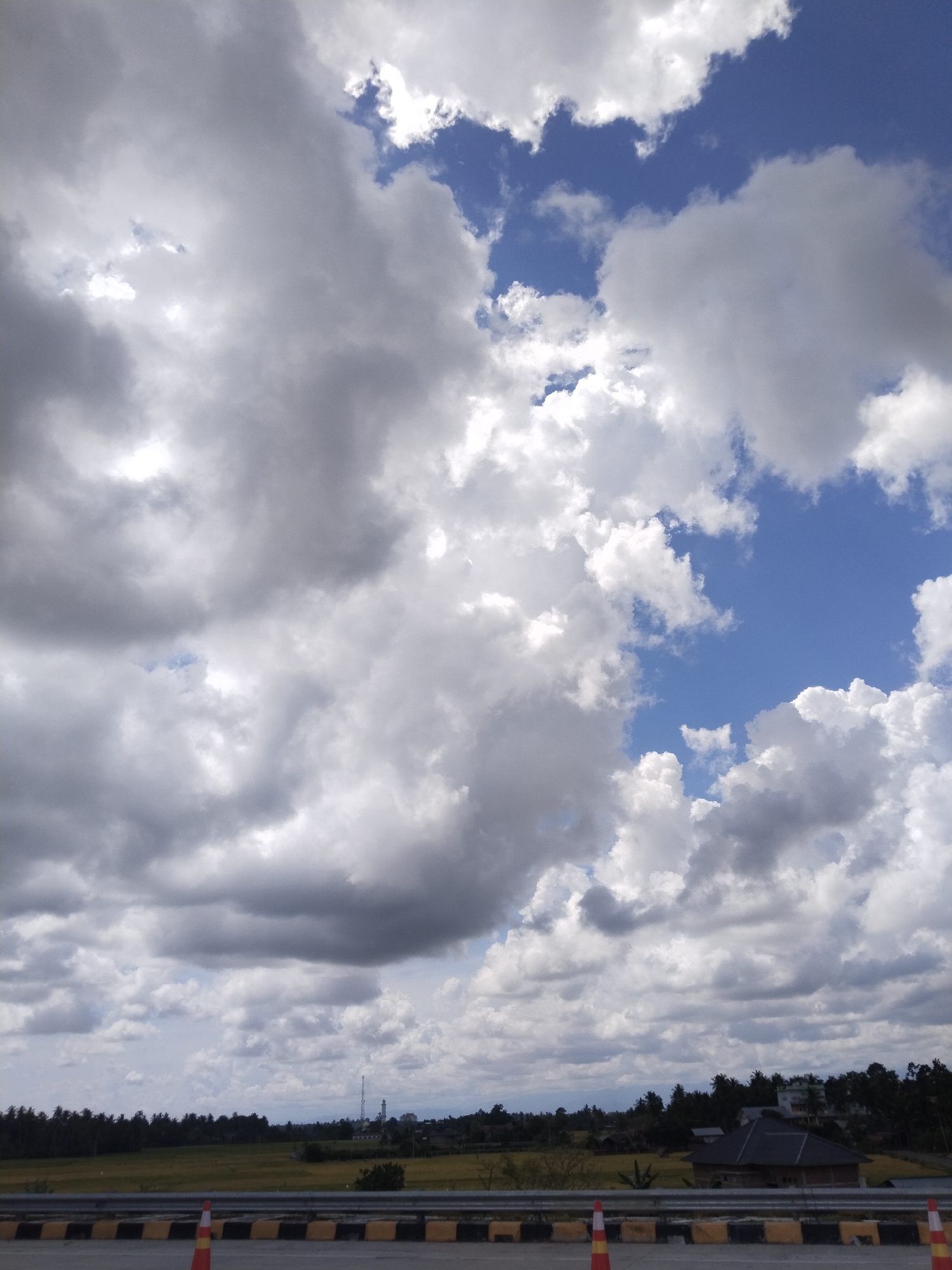 The image shows a cloudy sky over Capa Paloh, Indonesia. The sky is mostly covered by large, grey cumulus clouds, with patches of blue sky visible in between. Below the clouds is a flat landscape of rice fields, bordered by trees and some buildings. In the foreground is a section of a roadway, with a guardrail and traffic cones visible. The lighting suggests a partly cloudy day. A transmission tower is seen in the distance.