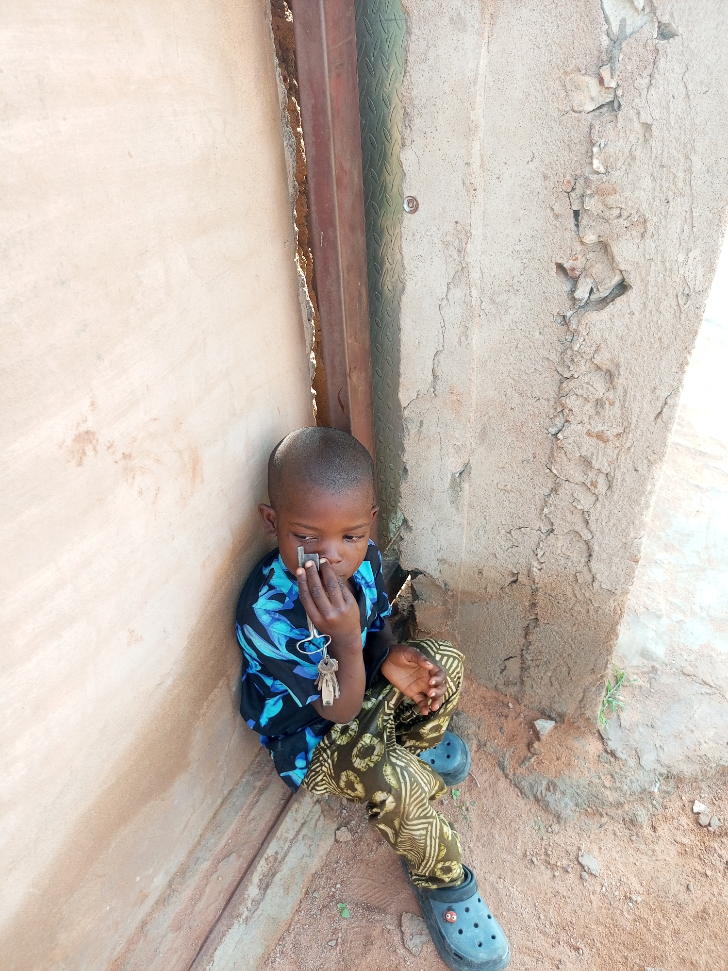 A young boy sits in a corner of a building in Tempure, Nigeria, leaning against a light-colored wall. He is wearing a black shirt with blue floral patterns and patterned pants. The boy is holding a set of keys in his right hand, bringing them to his mouth. His feet are clad in dark blue Croc-style shoes. The ground is covered in dirt and small stones, and to the right of the boy, a rough, textured concrete pillar stands. A metal doorway or frame is visible behind him, to the left. The environment appears to be an outdoor area, possibly a courtyard or an alleyway, with evidence of construction or decay in the surrounding structures.