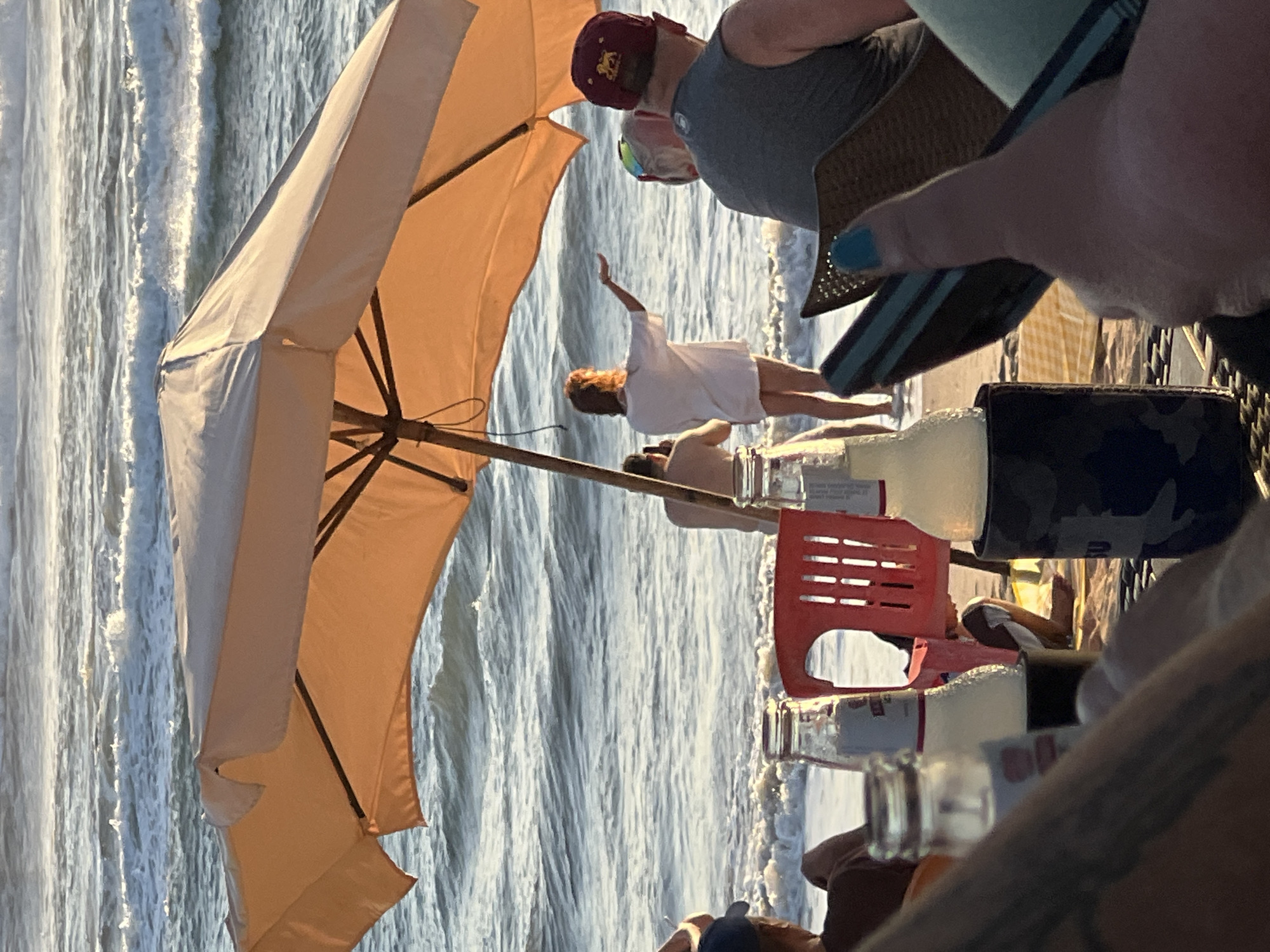 In Kuta, Bali, Indonesia, a group of people enjoy a sunset on the beach under a large, orange beach umbrella. The ocean is calm with gentle waves, and the sky is a warm, hazy golden color, indicating late afternoon or early evening. In the foreground, several bottles of drinks are visible, one in a black insulated koozie. A person's thumb with blue nail polish is in the lower right corner, giving a thumbs-up gesture. In the middle ground, a woman in a white t-shirt and shorts stands facing the ocean, with her arm extended as if reaching for something. Another person is visible behind her, taking a photo. To the right, a man wearing a gray tank top and a maroon baseball cap with a lion logo is seated. A red plastic chair is positioned near the water's edge. The overall scene suggests a relaxed and leisurely atmosphere on a tropical beach.
