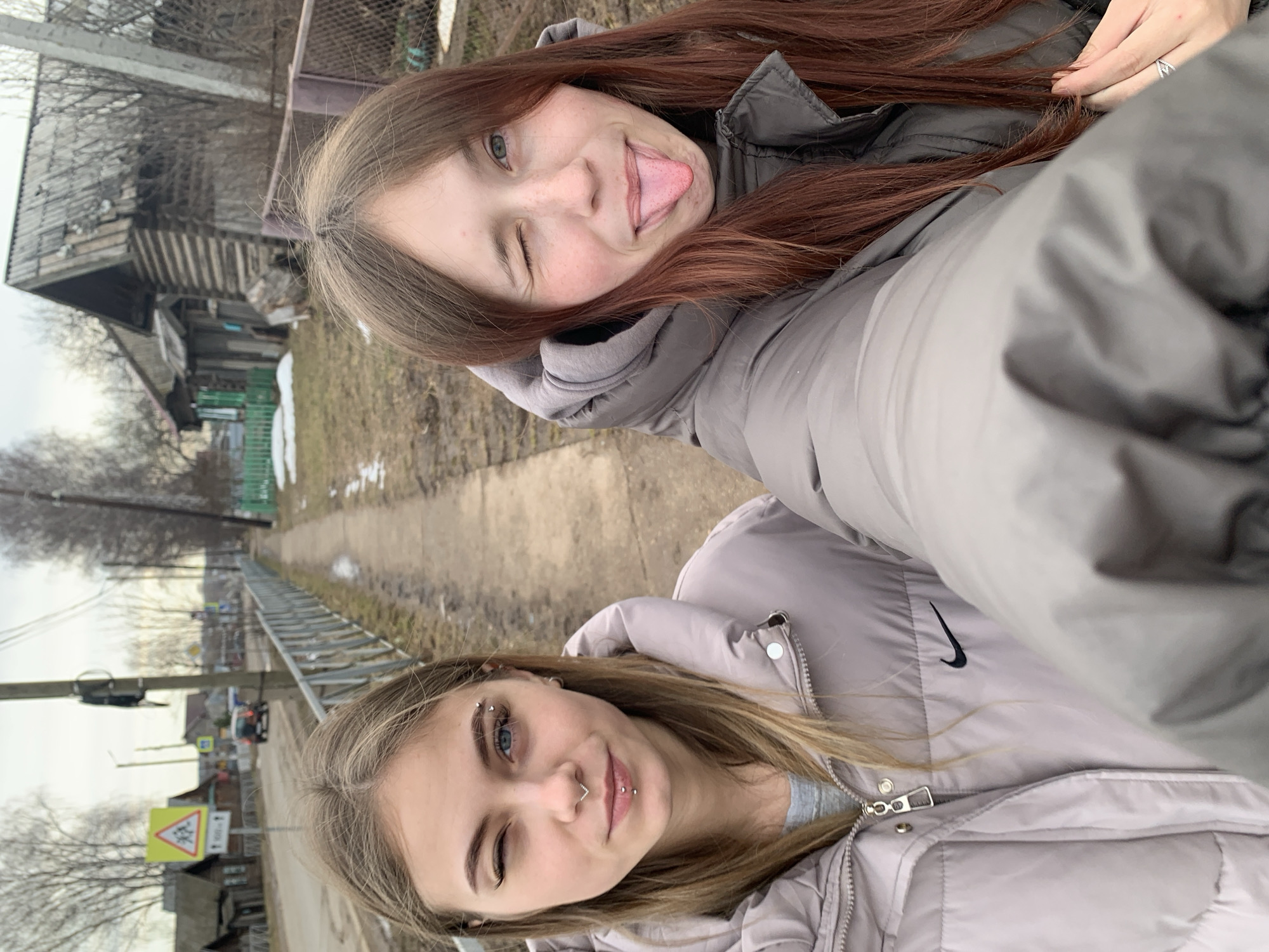 Two young women are posing closely for a selfie on a street in Фоки, Russia. Both are wearing winter jackets; one has long auburn hair and is sticking her tongue out while winking, while the other has blond hair, piercings on her eyebrow, nose, and lip, and is also winking. In the background is a sidewalk running along the edge of a bare field. A low metal fence parallels the sidewalk, separating it from the field. On the other side of the sidewalk are a utility pole, road signs, and a view of rural buildings. The weather appears overcast.