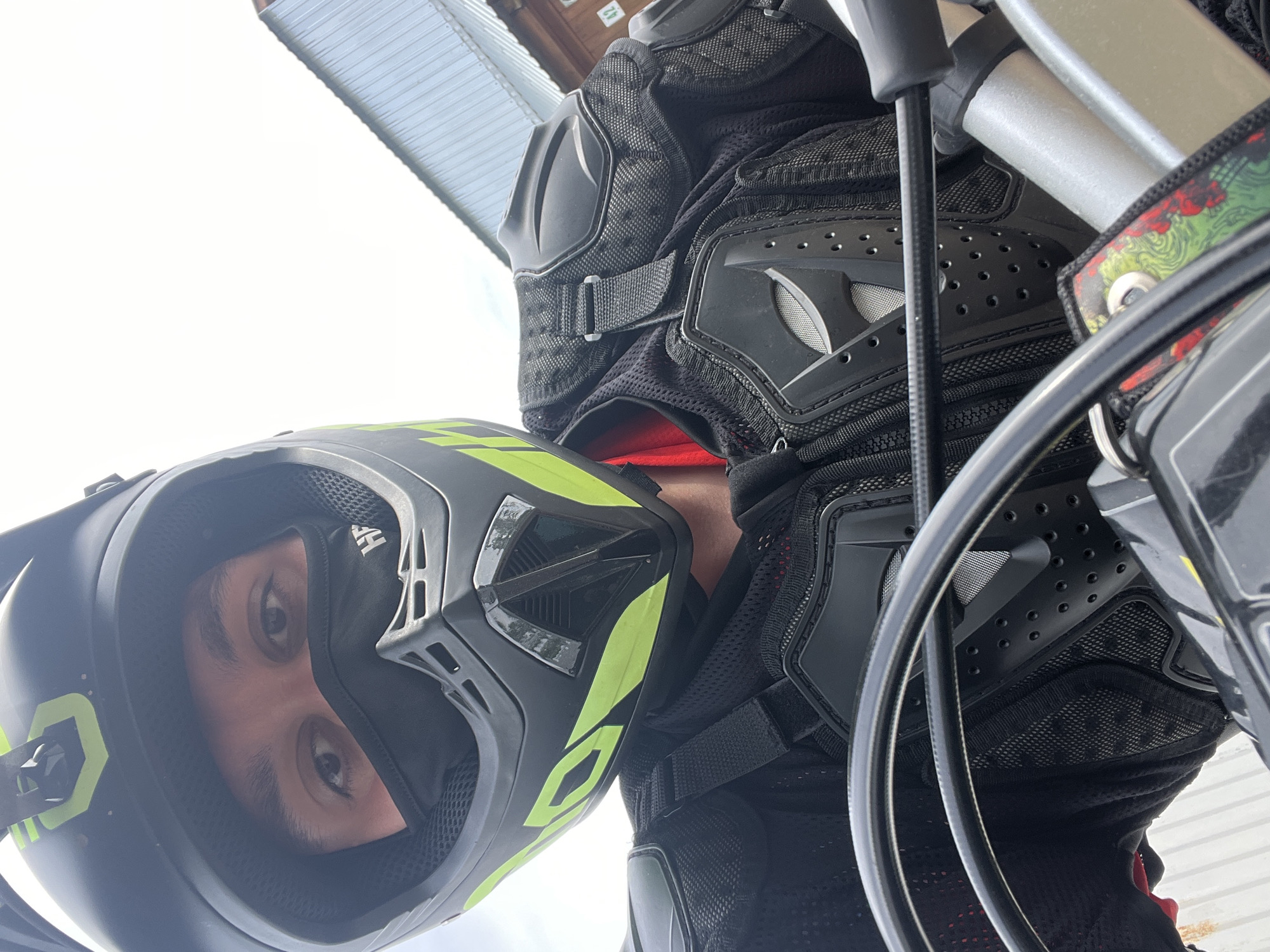 The image shows a person wearing a black and neon green motocross helmet and black protective gear, likely chest and shoulder armor. The person is looking directly at the camera. In the background, a building with a corrugated metal roof is visible. The location is stated as Vasilyevo, Russia, though there is no visual confirmation within the image itself.