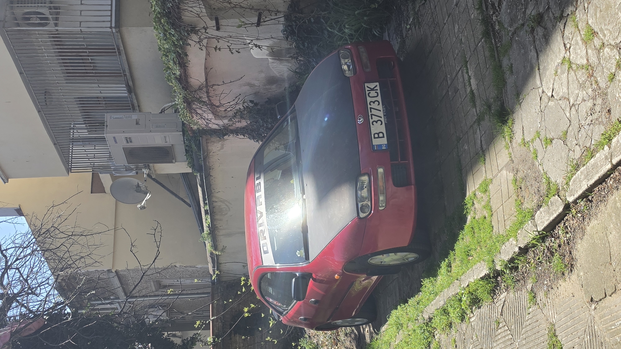 The image depicts a red Mazda parked on a narrow street with cracked concrete pavement and grass growing between the cracks. The car has a black hood and a license plate reading "B 3773 CK". A white sticker with "DRIVABL" is visible on the windshield. The surrounding environment includes buildings with air conditioning units and satellite dishes, as well as bare trees and greenery. The location is likely a residential area within the city of Varna, Bulgaria, based on the license plate. No people are visible in the image.