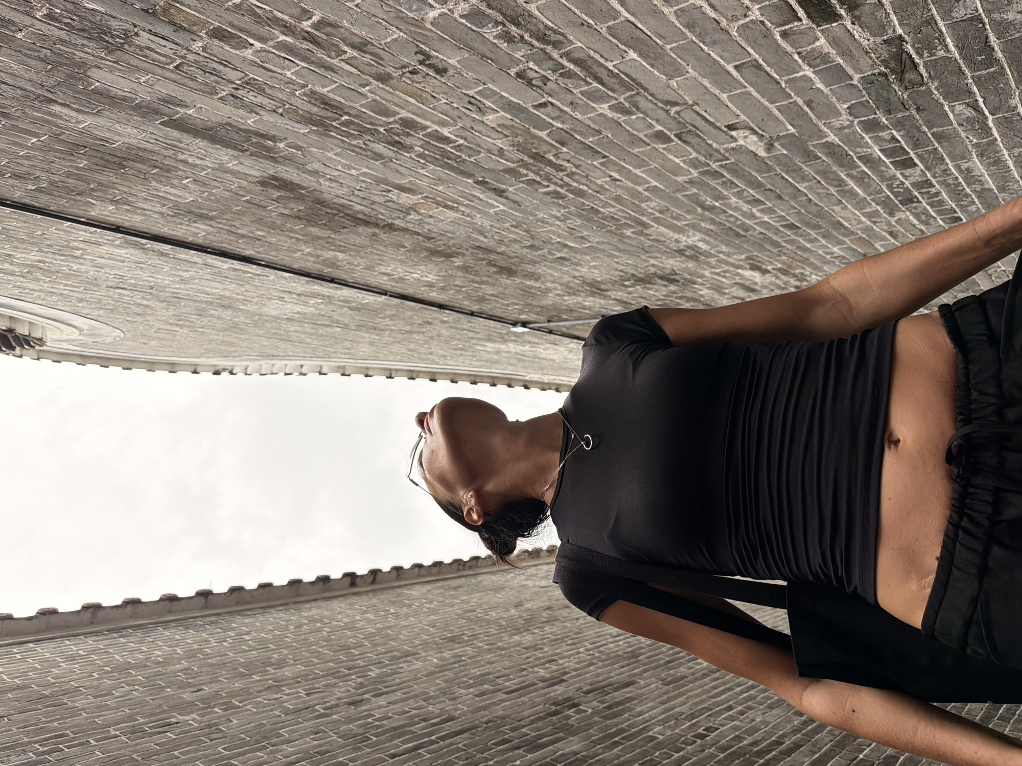 A low-angle medium shot captures a woman standing in a narrow alleyway in Chancheng District, China. She is wearing a black, fitted t-shirt that exposes her midriff and black drawstring pants. The woman is looking upwards with a slight tilt of her head, wearing thin-rimmed glasses and a necklace. A black tote bag is slung over her left shoulder. The alley walls, made of gray brick, rise steeply on either side, converging towards a pale, overcast sky. The overall scene suggests an urban exploration or casual outing in a historic part of the city.