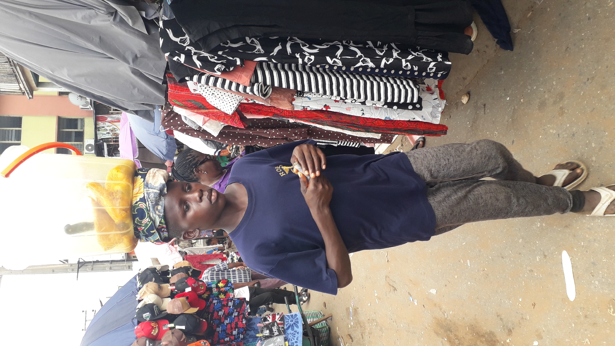 The image depicts a scene at a market in Nkpor, Nigeria. A young man wearing a blue t-shirt and grey sweatpants stands on a concrete floor, holding small objects in his hands. He also wears light-colored sandals. He is positioned amidst market stalls.
Behind him are stacks of clothing items, including patterns such as black and white swirls, stripes, polka dots, and floral prints. To his left is a clear plastic container holding orange-colored food items, carried atop someone's head with a patterned head wrap. A woman in purple is visible behind him and the container. Further left are caps and umbrellas.
The background reveals buildings, suggesting an urban setting. The ground shows signs of wear and tear.