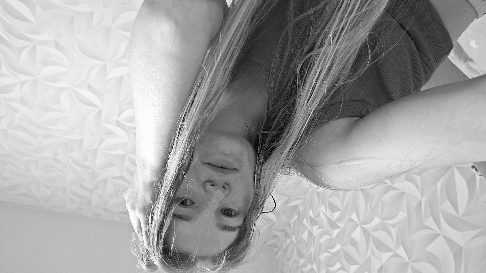 This is a grayscale, close-up, low-angle selfie of a young Caucasian woman with long, light-colored hair and freckles. She is looking directly at the camera, with her left arm bent and her hand resting on the back of her head. She is wearing a ribbed top. The background is a wall with a geometric, textured pattern. The scene appears to be indoors. No specific location in Chaikovsky, Russia can be identified from the image.