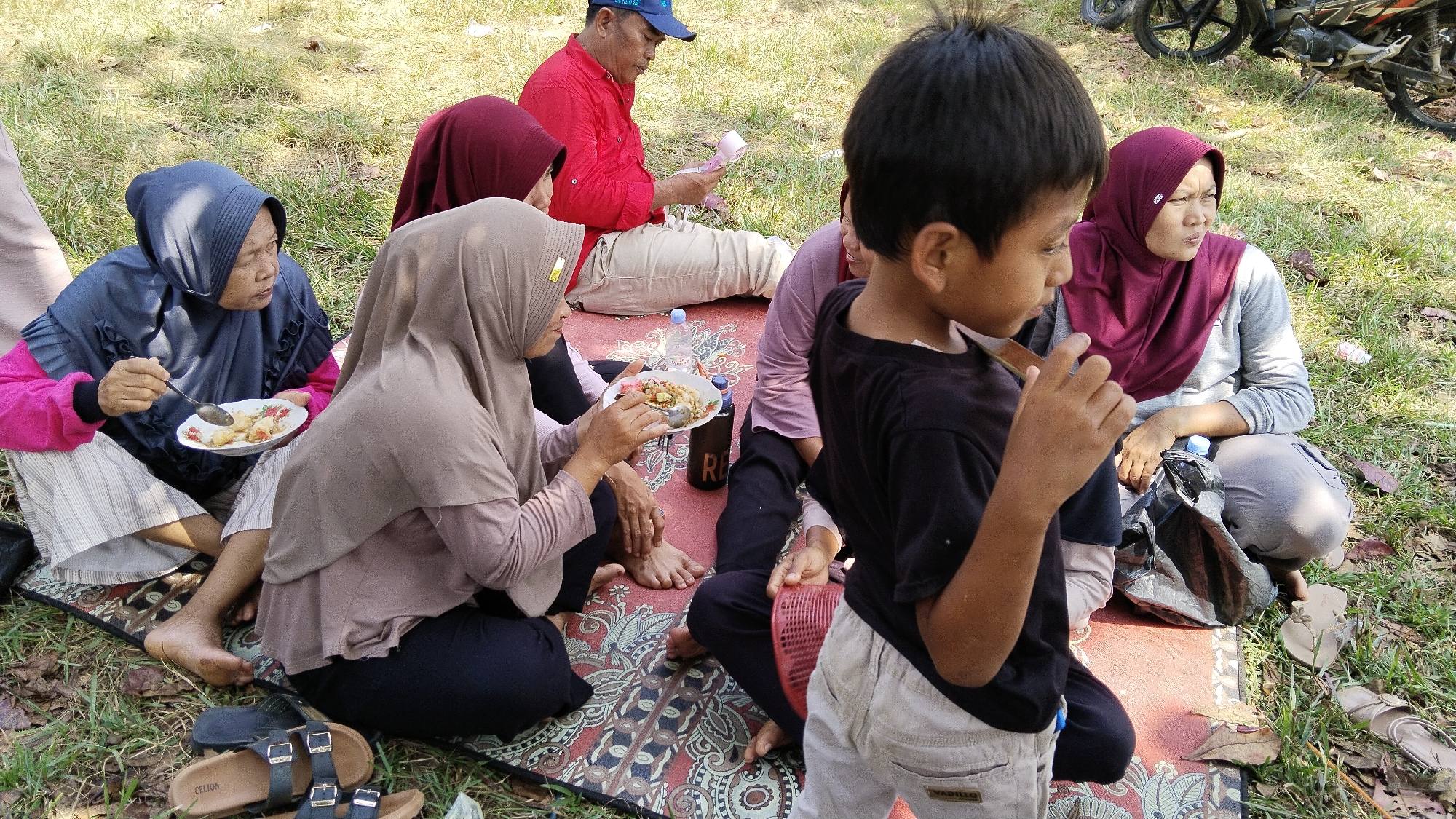 In Margatunggal, Indonesia, a group of people are having a picnic on a patterned mat spread on a grassy area. A young boy in a black t-shirt and light-colored shorts stands in the foreground, looking to his right. To his left, a woman wearing a blue hijab and a pink long-sleeved shirt is eating from a plate with a spoon. Beside her, another woman in a beige hijab is also holding a plate. In the background, a man in a red shirt and blue cap is seated, appearing to be looking at something in his hands. Another woman in a maroon hijab is seated to his right. The environment is outdoors, with dry leaves scattered on the grass, and the sun is casting shadows. Part of a motorcycle is visible in the upper right corner of the image.