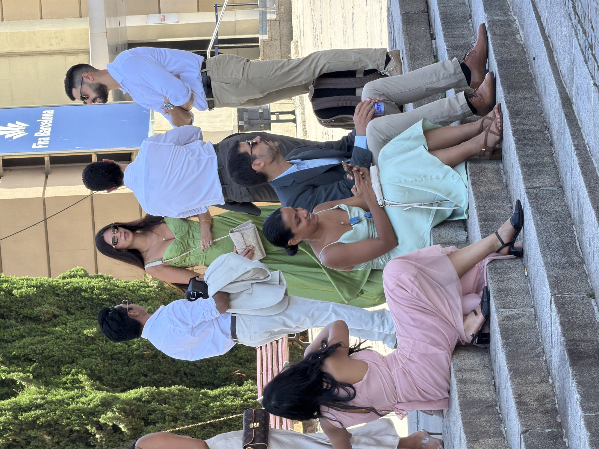 This image depicts a group of people gathered outside the Fira Barcelona exhibition center, as indicated by signage in the background. The scene appears to be a public outdoor space with stone steps leading up to a building.
In the foreground, two women are seated on the steps. The woman on the left is wearing a pale pink, floor-length dress and black heeled sandals. She is looking downwards, and her back is mostly to the camera. The woman on the right is wearing a pale green, knee-length dress and brown strappy heeled sandals. She is looking at her phone, which she holds in her hands. Beside her, a man in a dark suit and light blue shirt is seated, also looking down. He is wearing beige trousers and brown dress shoes.
Further up the steps, several individuals are standing. A man in a white shirt and beige trousers is standing with his arms crossed, looking to his right. Another man in a white shirt and beige trousers is facing away from the camera, holding a light-colored jacket over his arm. A woman in a lime green dress is standing between them, holding a light-colored handbag.
The environment includes lush green trees to the left and the facade of the Fira Barcelona building to the right. The overall atmosphere suggests a social gathering, possibly an event or a break during an exhibition.