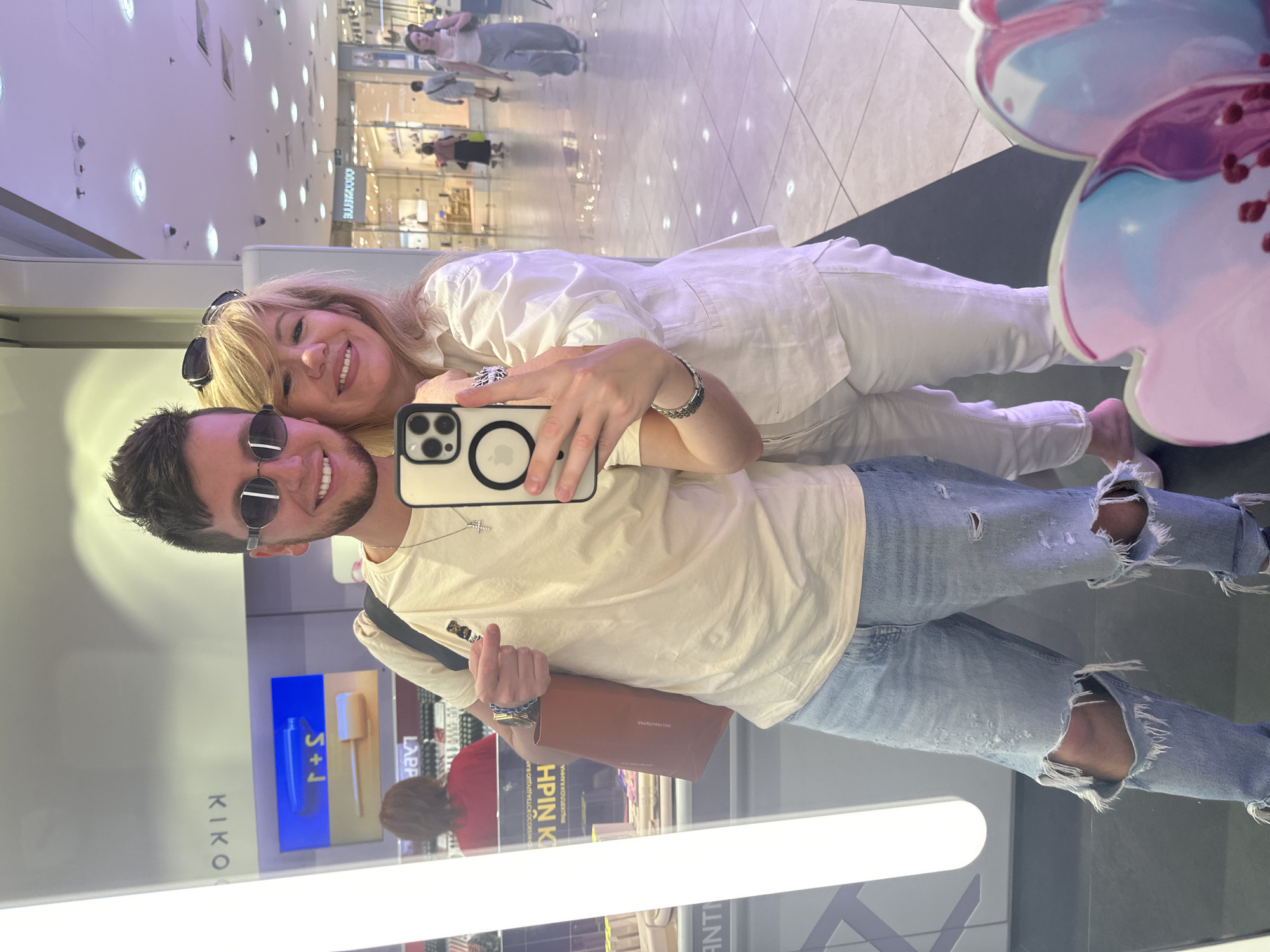 A young man and an older woman take a selfie in a brightly lit indoor shopping mall. The man, on the left, wears ripped light-wash jeans, a pale yellow t-shirt with a small bear graphic, and oval sunglasses. He holds a brown paper bag in his left hand. The woman, on the right, wears a white linen shirt and white pants. Both are smiling. In the background, various shops and shoppers are visible. A large, brightly lit vertical display is to the left of the pair, reflecting in a mirror, obscuring part of the scene.
