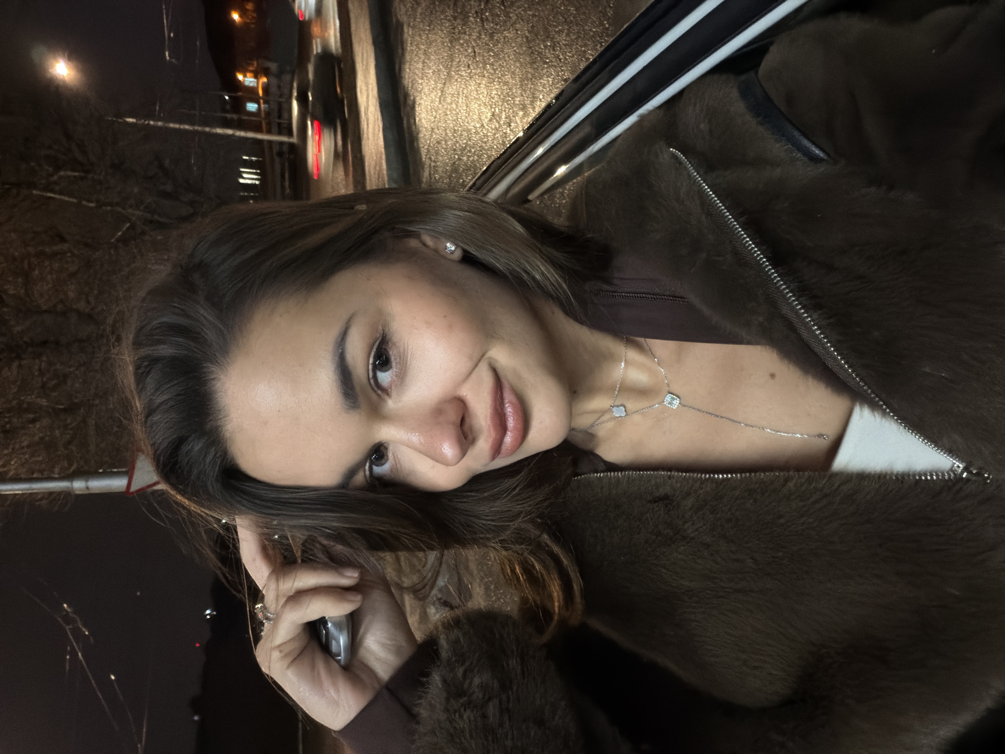 A woman is in the back of a car, possibly in the city of Krasnoyarsk, Russia, during a night scene with city lights visible in the background. She is fair-skinned with brown hair, wearing a brown fur coat over what appears to be a brown top and a white garment underneath. She is also wearing a silver necklace with square pendants. She is holding a phone in her left hand. The car's window frame is visible behind her. The background shows blurred city lights, suggesting movement.
