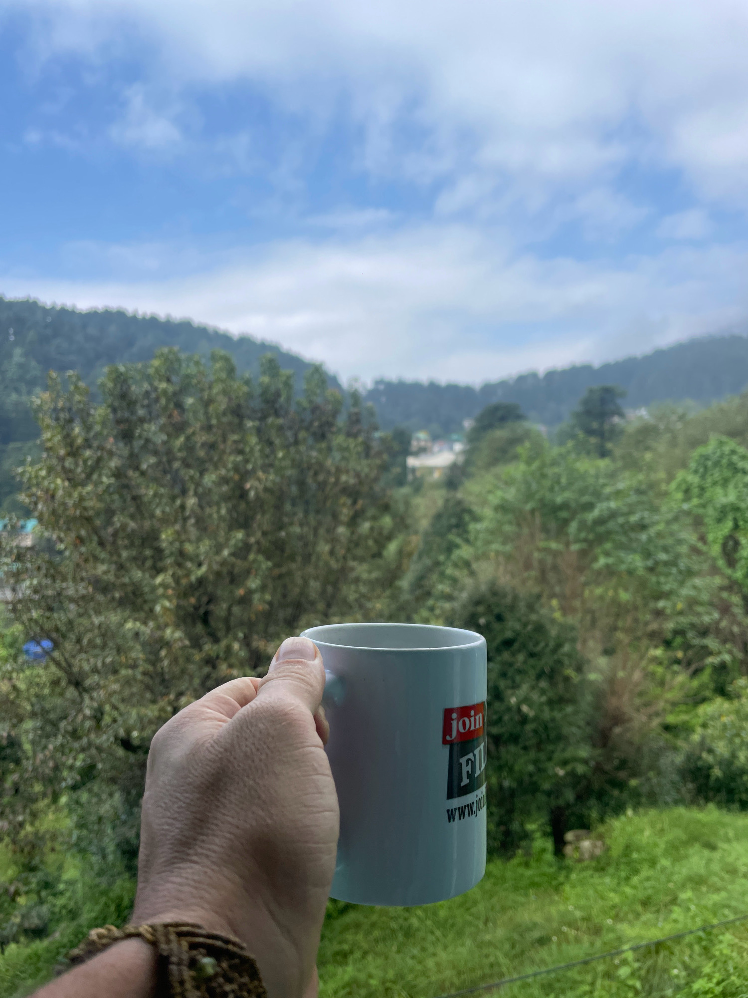 A hand holding a white coffee mug with the logo "JOIN FIL" and "www.jo" is in the foreground. The mug is held against a backdrop of lush green trees and hills under a cloudy blue sky. The scene appears to be in Bhagsu, India, with the natural landscape of the Himalayas evident. The hand is wearing a brown woven bracelet.