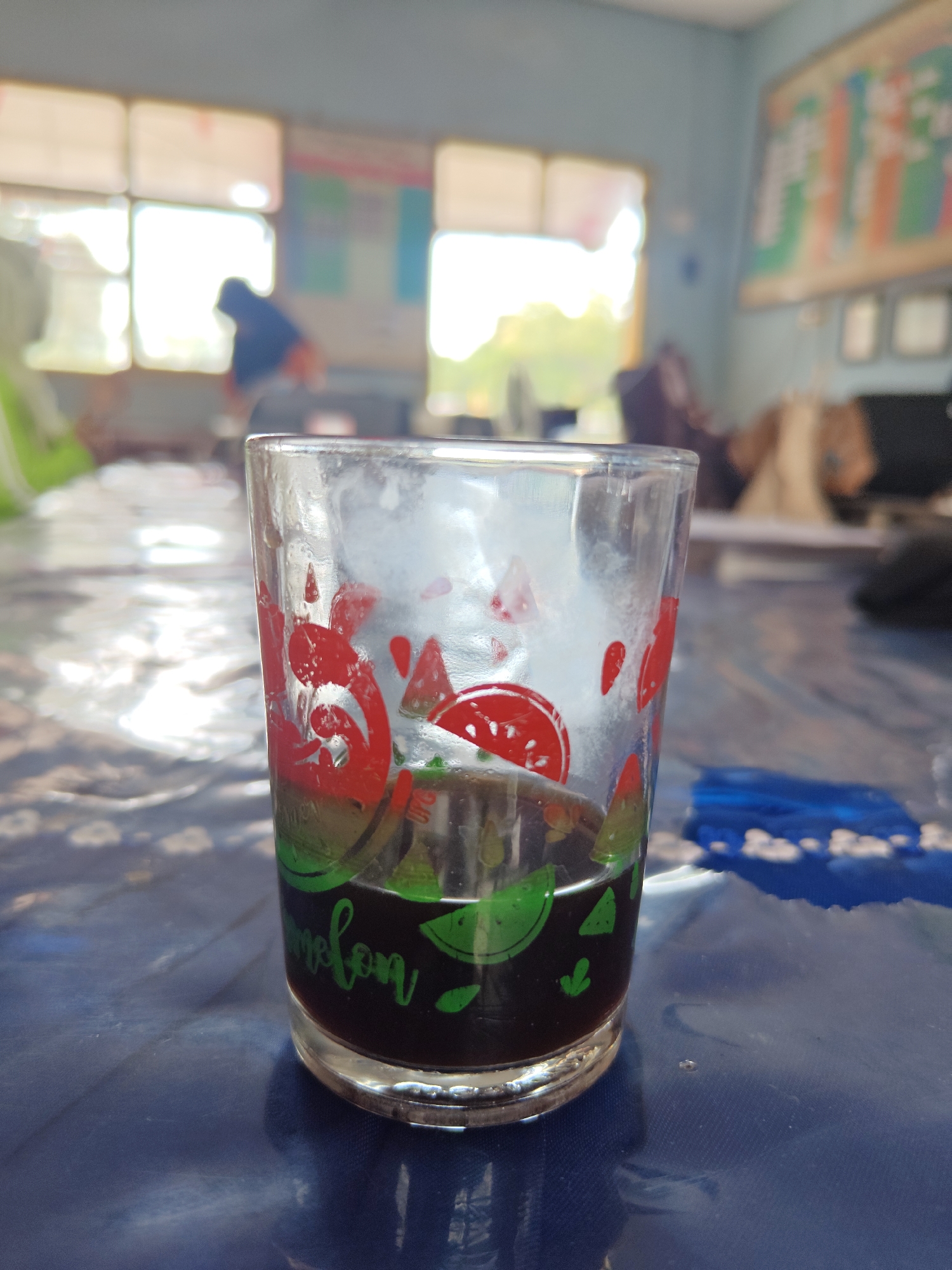 A close-up shot focuses on a clear glass with red and green watermelon designs, containing a dark liquid, likely coffee or tea. The glass rests on a blue, patterned surface, possibly a tablecloth. In the background, through windows, sunlight illuminates a room, with blurred figures and posters on the wall, suggesting an indoor environment like a classroom or meeting hall in Cermee, Indonesia. The overall scene is casual and likely domestic or educational.